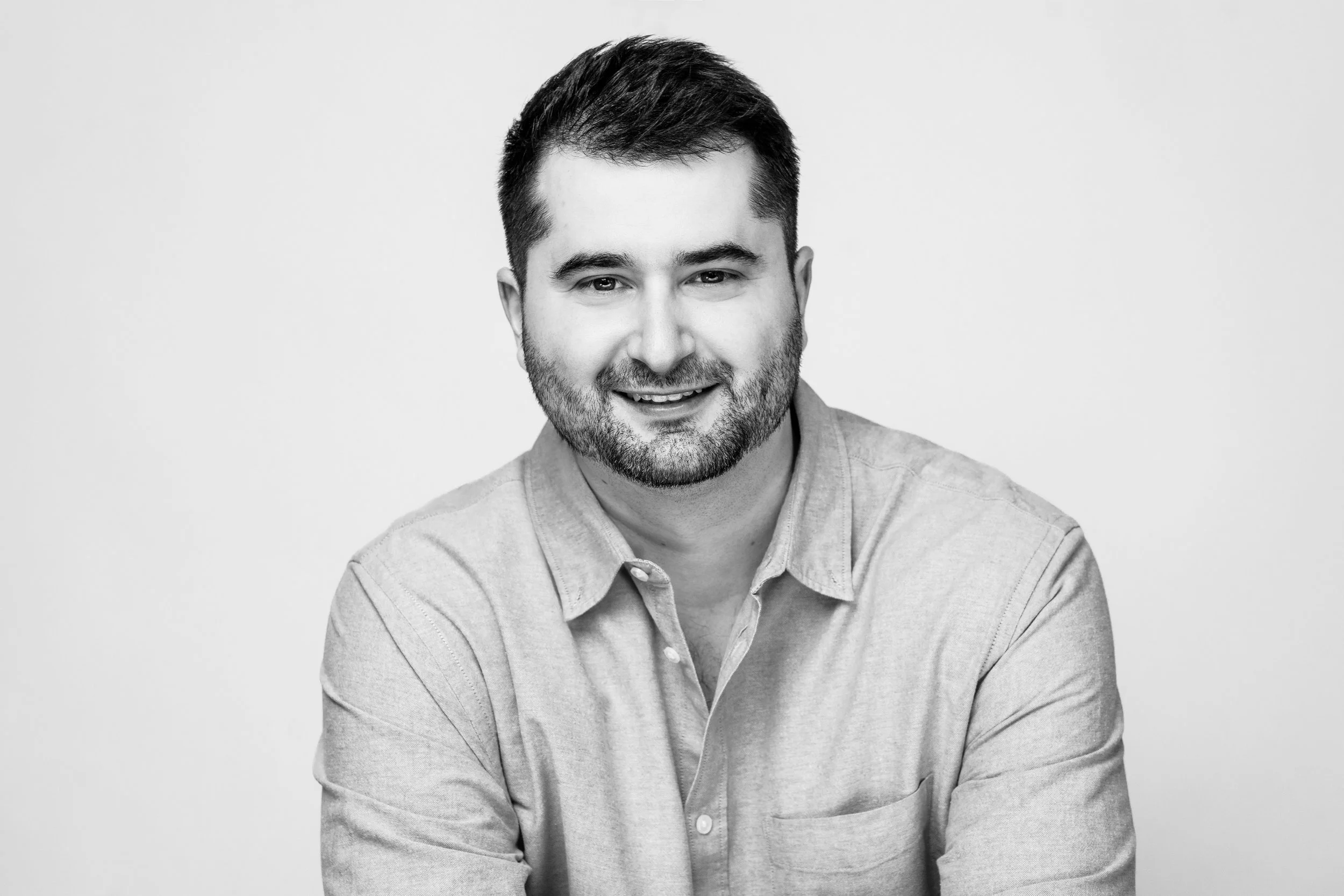 Black and white portrait of a smiling man with short dark hair, beard, and mustache, wearing a light-colored button-up shirt with rolled-up sleeves, against a plain light background.