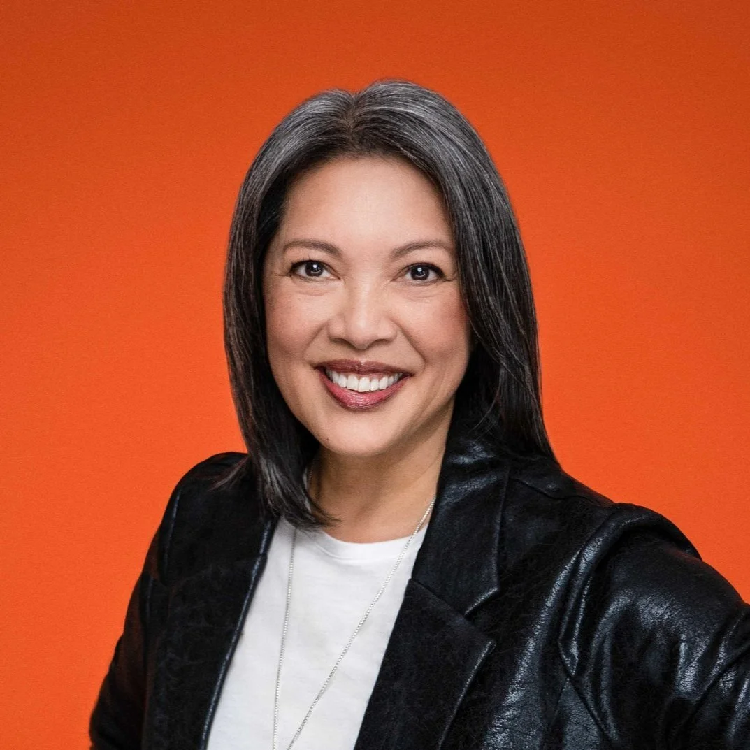 A woman with shoulder-length black hair smiling in front of an orange background, wearing a black leather jacket and a white shirt.