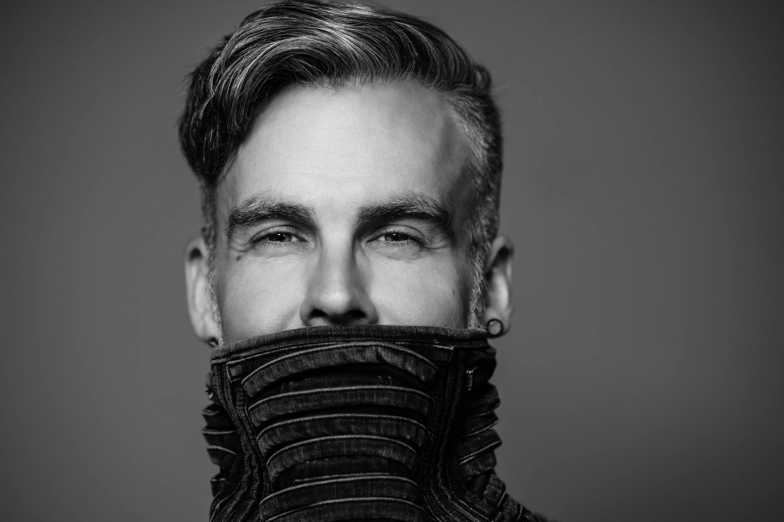 Black and white portrait of a person with short hair, partially covering their face with a high-collared, textured garment. Only their eyes and forehead are visible.