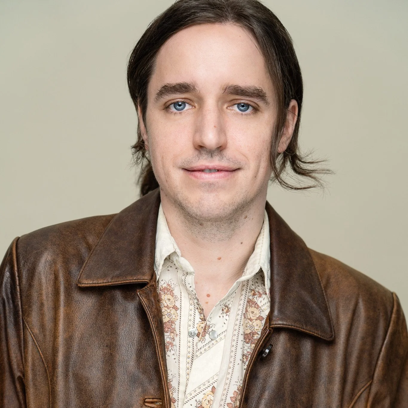 A young man with shoulder-length brown hair and blue eyes, wearing a brown leather jacket over a cream-colored shirt with a floral pattern, standing against a neutral background.