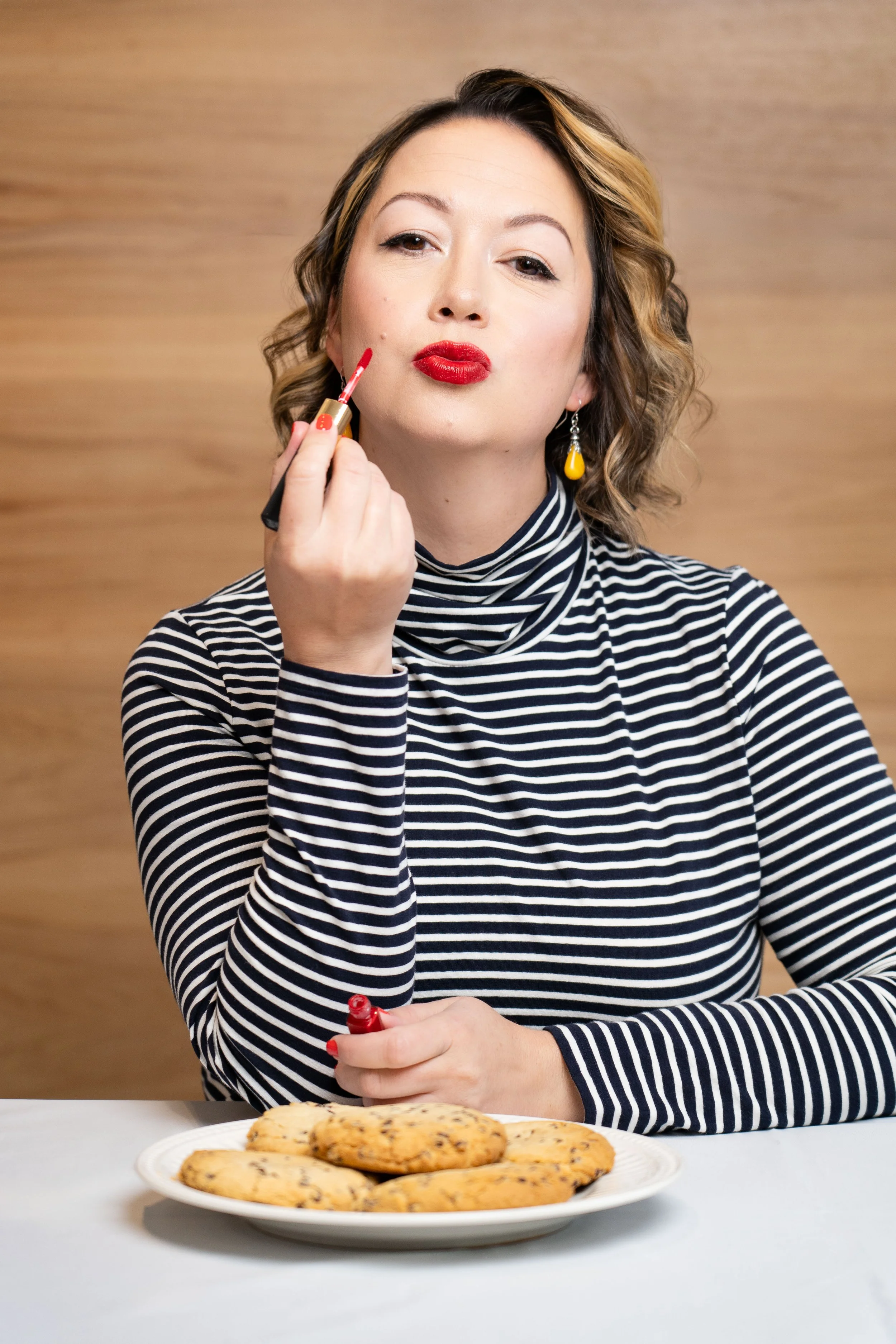 A woman with curly hair in a striped turtleneck applying red lipstick, sitting at a table with a plate of chocolate chip cookies.