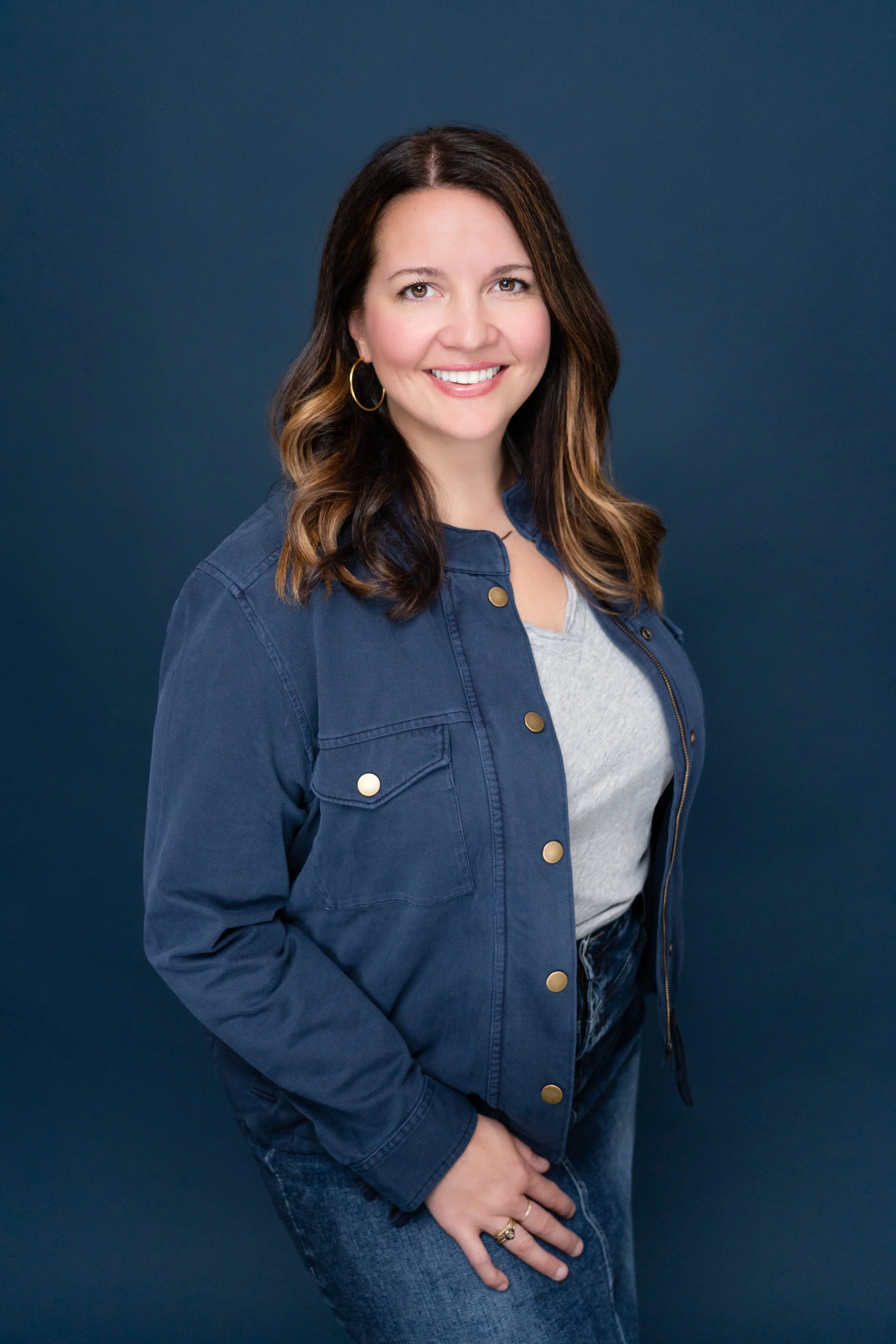 A woman with shoulder-length brown hair and light skin, smiling, wearing a gray t-shirt, a dark blue jacket, and blue jeans, standing against a dark blue background.
