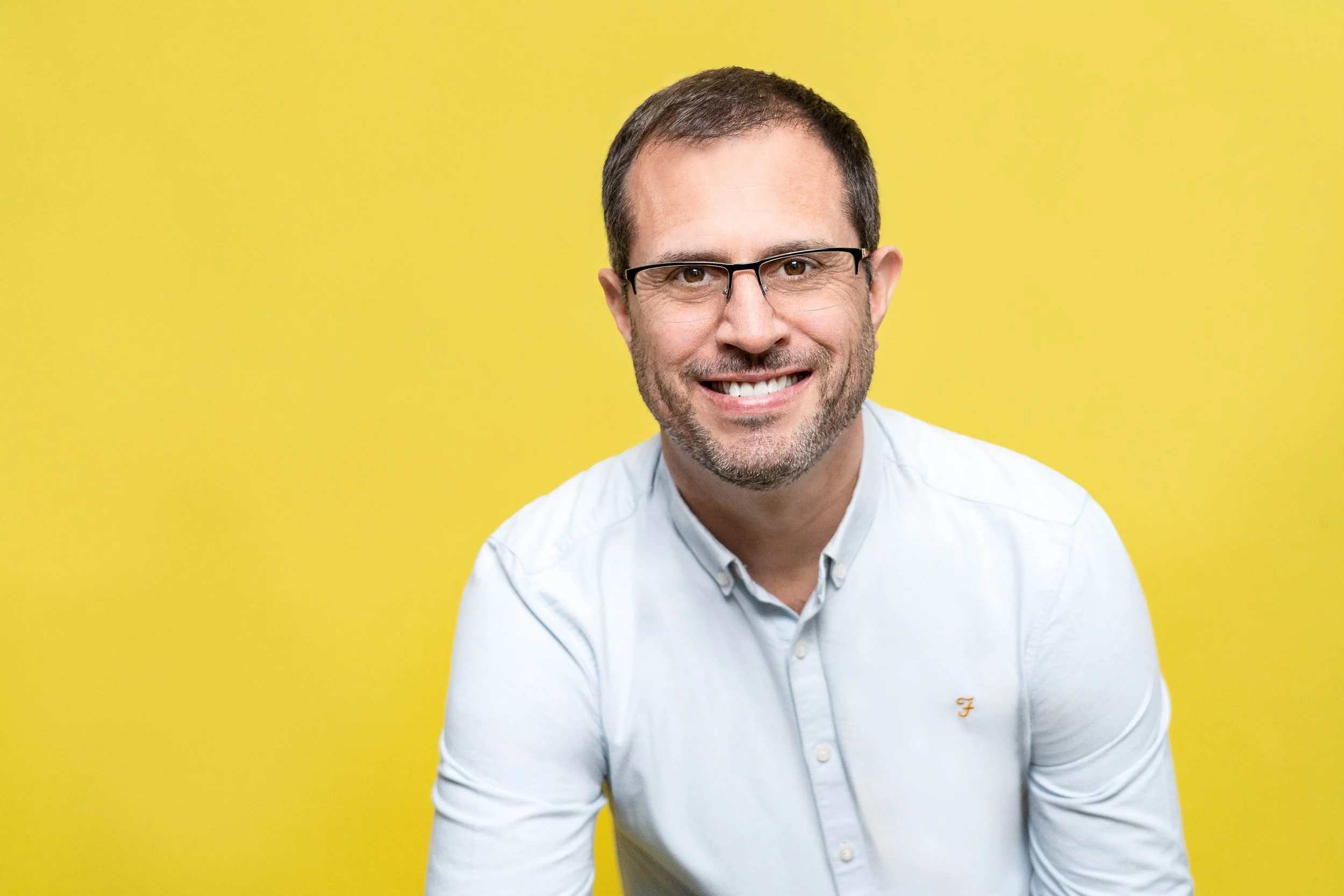 A smiling man with glasses and a beard wearing a white button-up shirt against a yellow background.