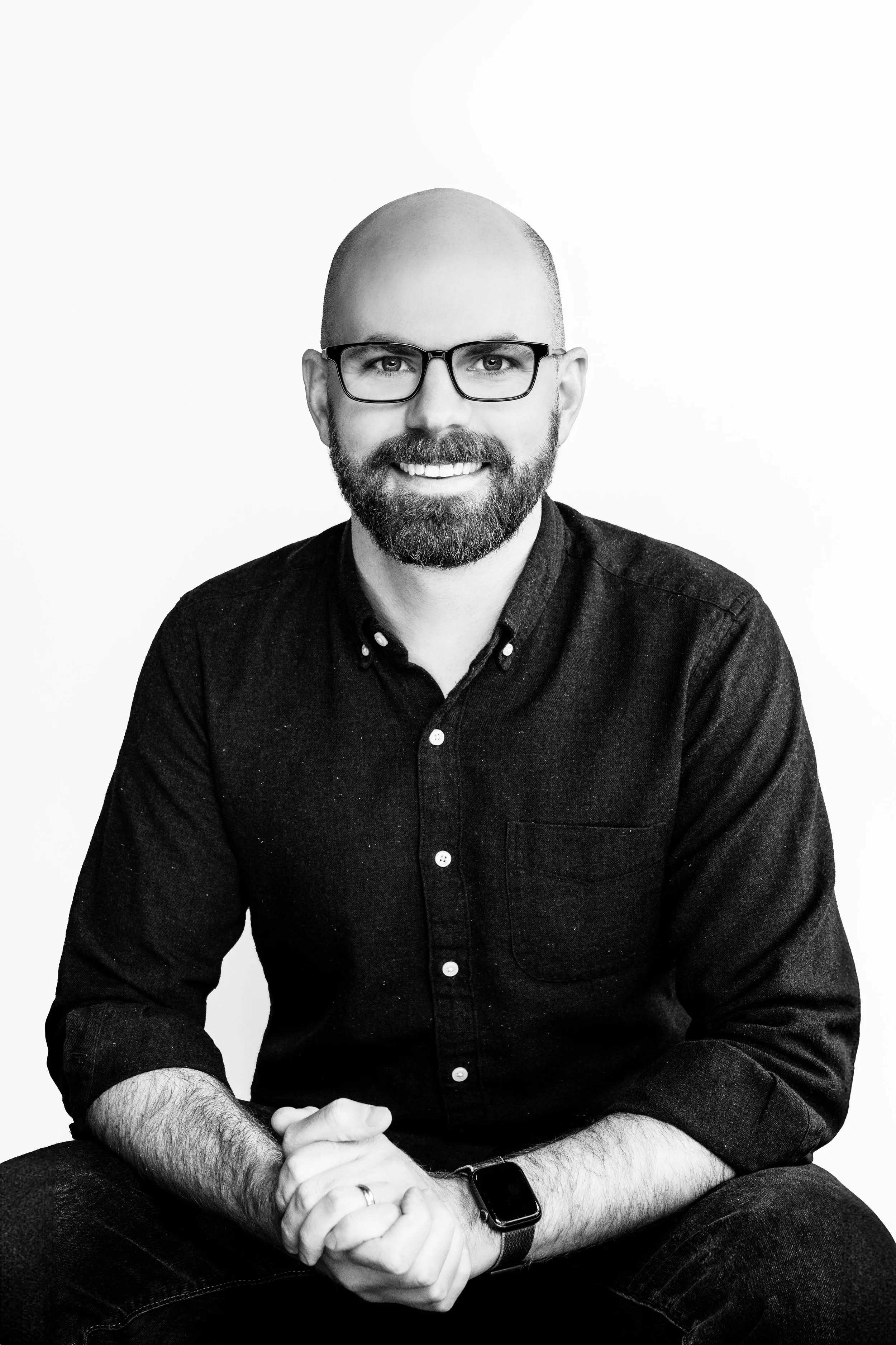 Black and white portrait of a smiling man with glasses, a beard, wearing a dark button-up shirt, sitting with hands clasped, against a plain background.