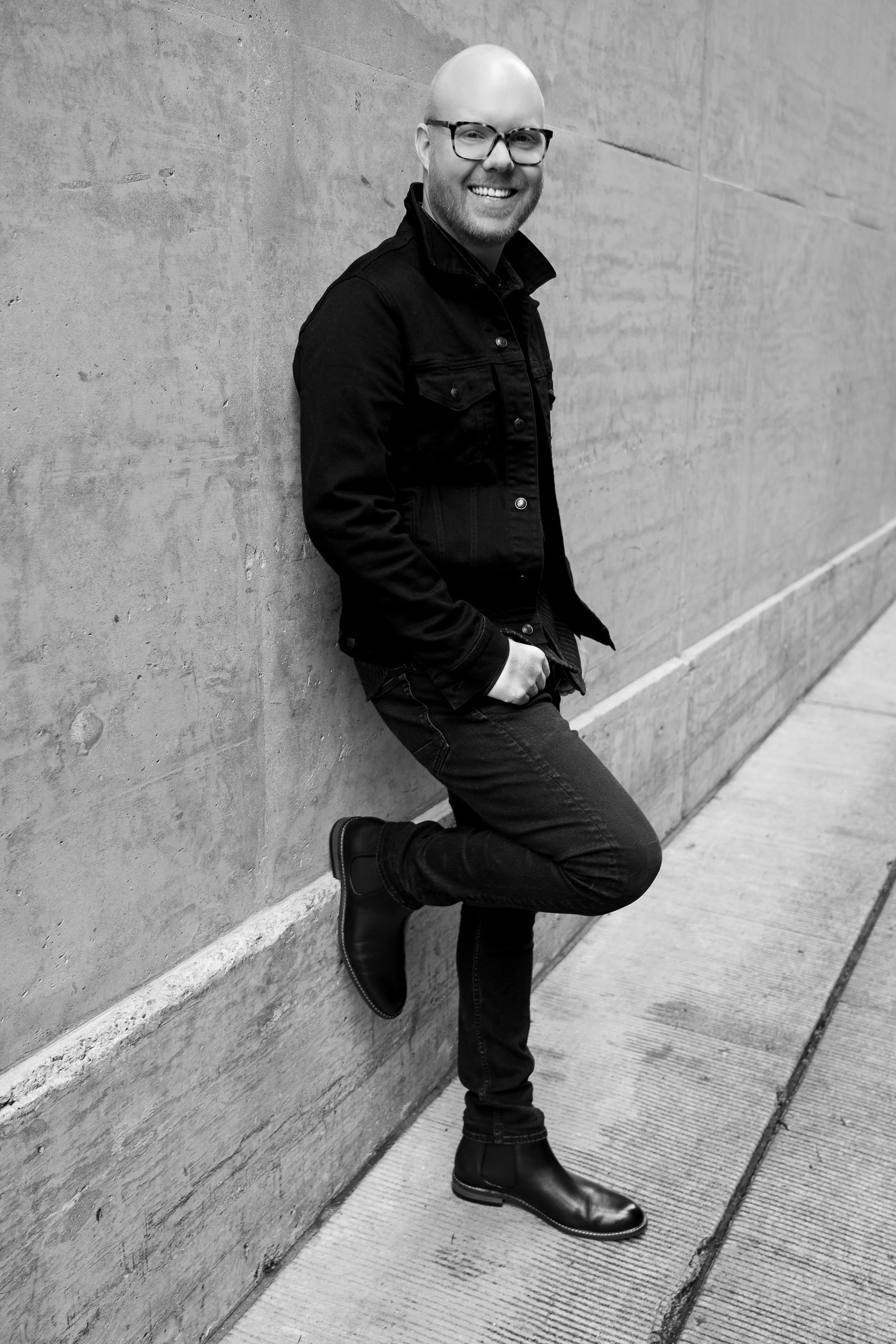 Black and white photo of a smiling man with glasses, bald head, wearing a black jacket, jeans, and boots, leaning against a concrete wall with one foot up against the wall.