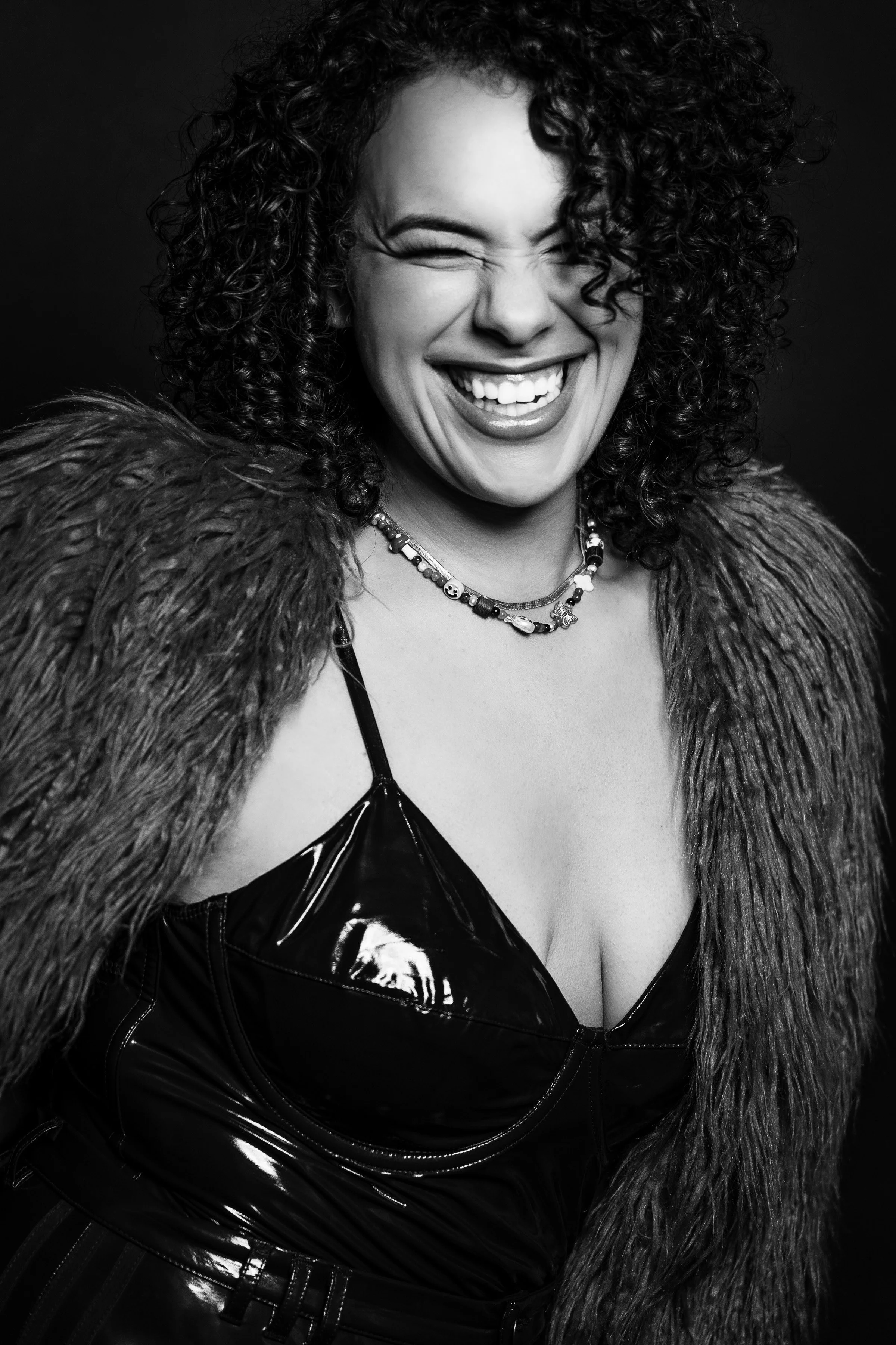 Black and white photograph of a woman with curly hair, smiling and winking, wearing a shiny leather top, a necklace, and a faux fur coat draped over her shoulders.