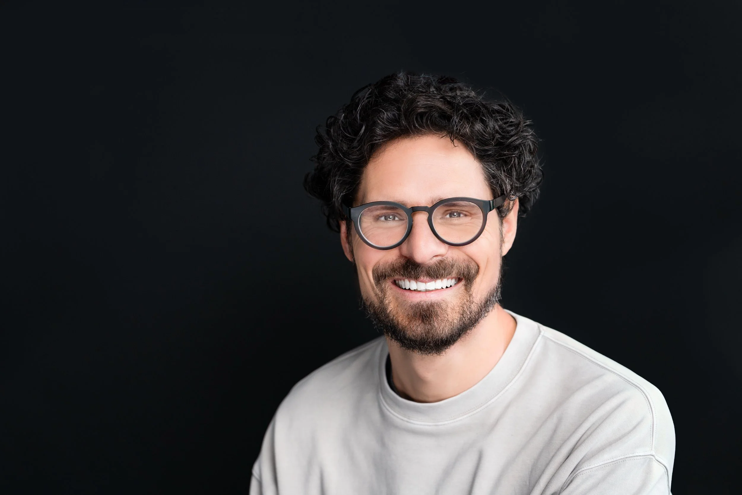 Smiling man with curly hair, glasses, and a beard, wearing a light-colored shirt, against a black background.