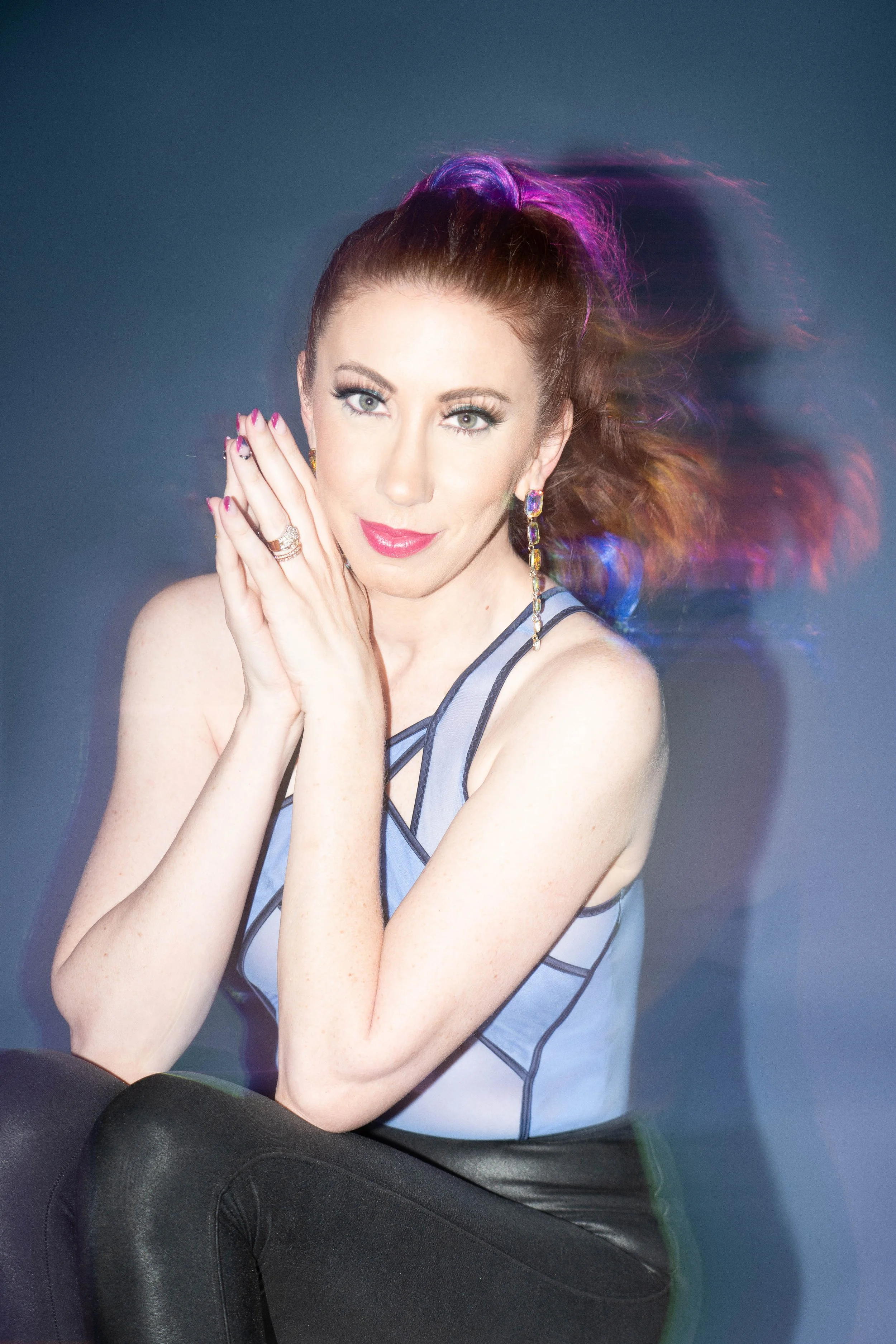 A woman with red hair, styled in a high ponytail with purple highlights, looking at the camera with her hands clasped near her face, wearing a sleeveless top and earrings, against a dark gray background with rainbow-colored motion blur.