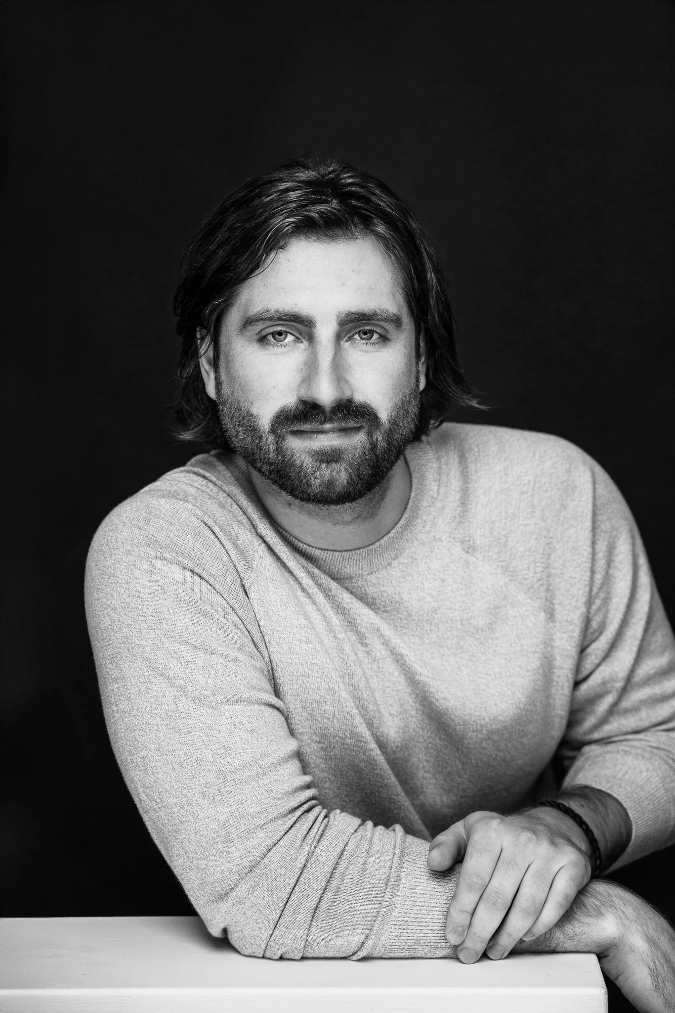 Black and white portrait of a man with dark, shoulder-length hair and a beard, wearing a light-colored sweater, sitting with arms crossed on a light-colored surface against a dark background.