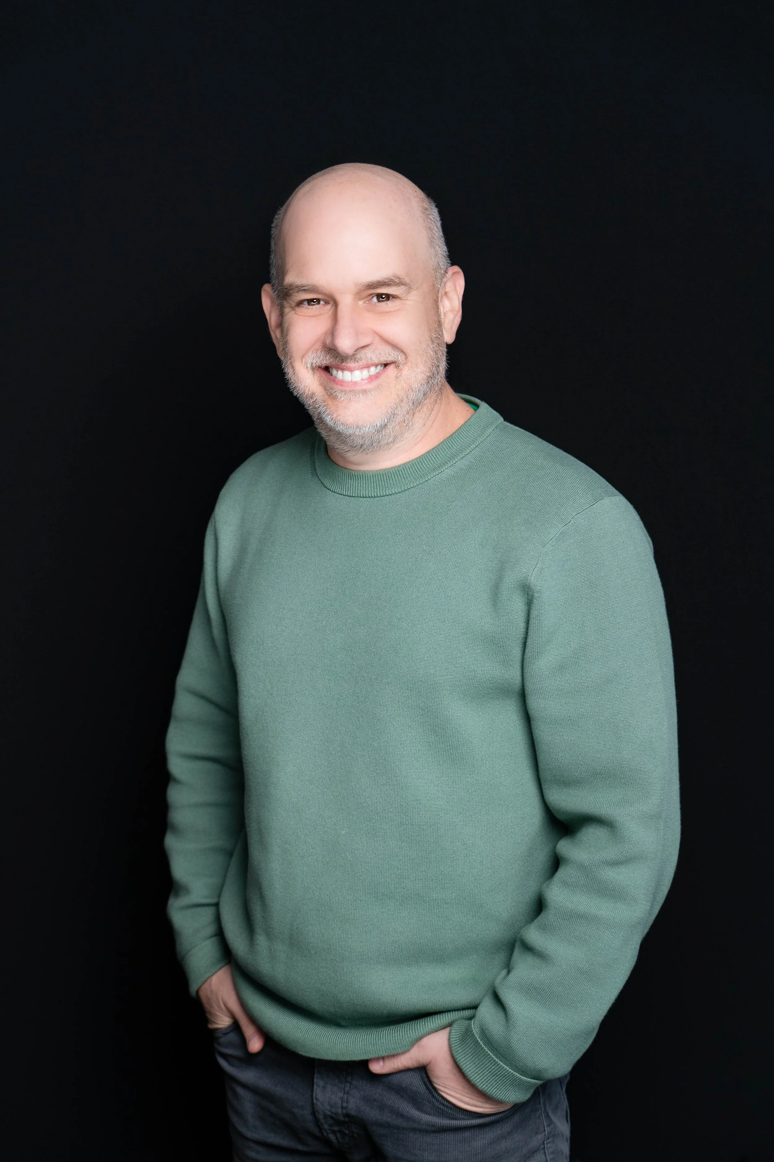 A middle-aged man with a bald head and gray beard wearing a green sweater, smiling and posing against a black background.