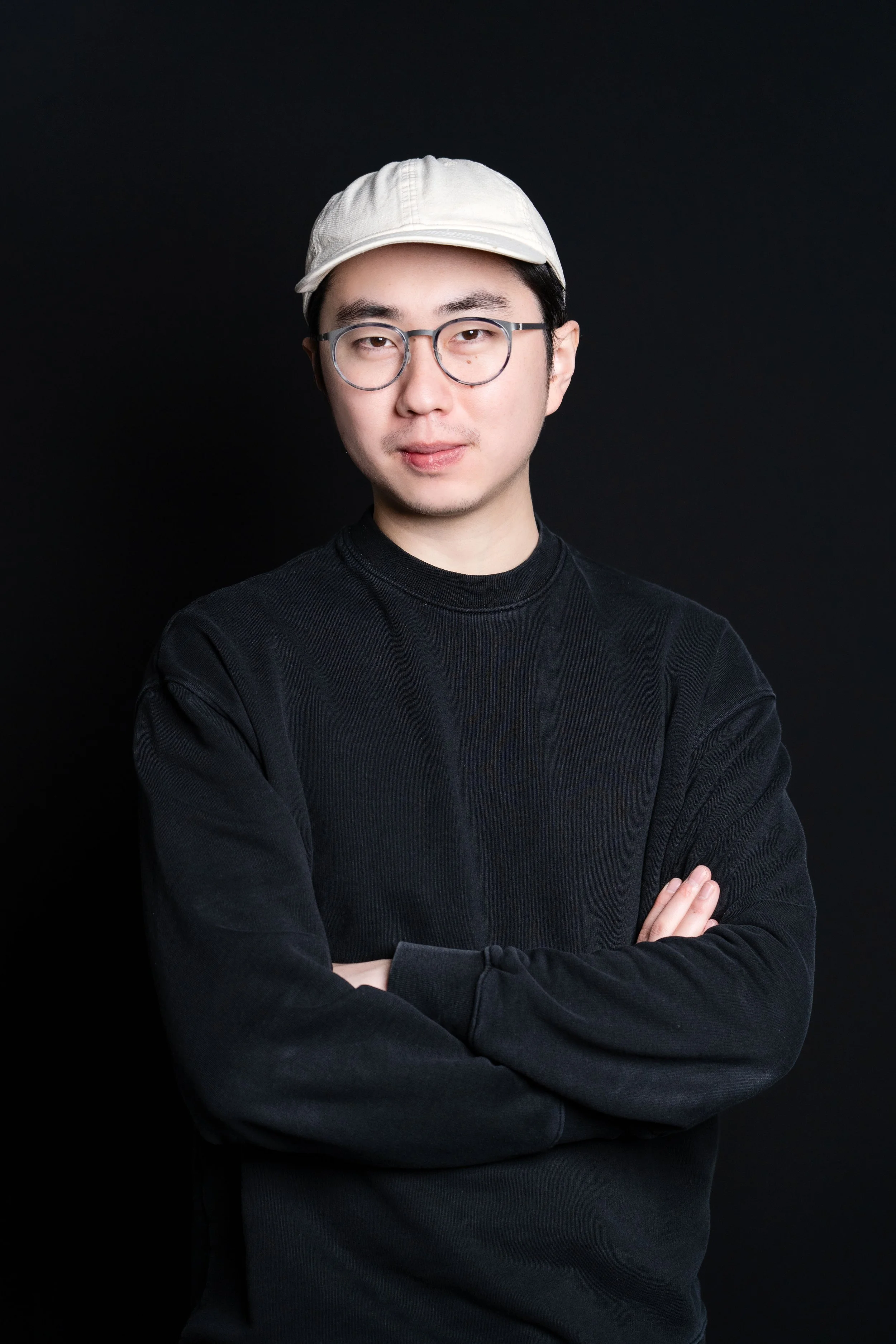 Young man wearing glasses, a light-colored cap, and a black long-sleeve shirt, standing with arms crossed against a black background.