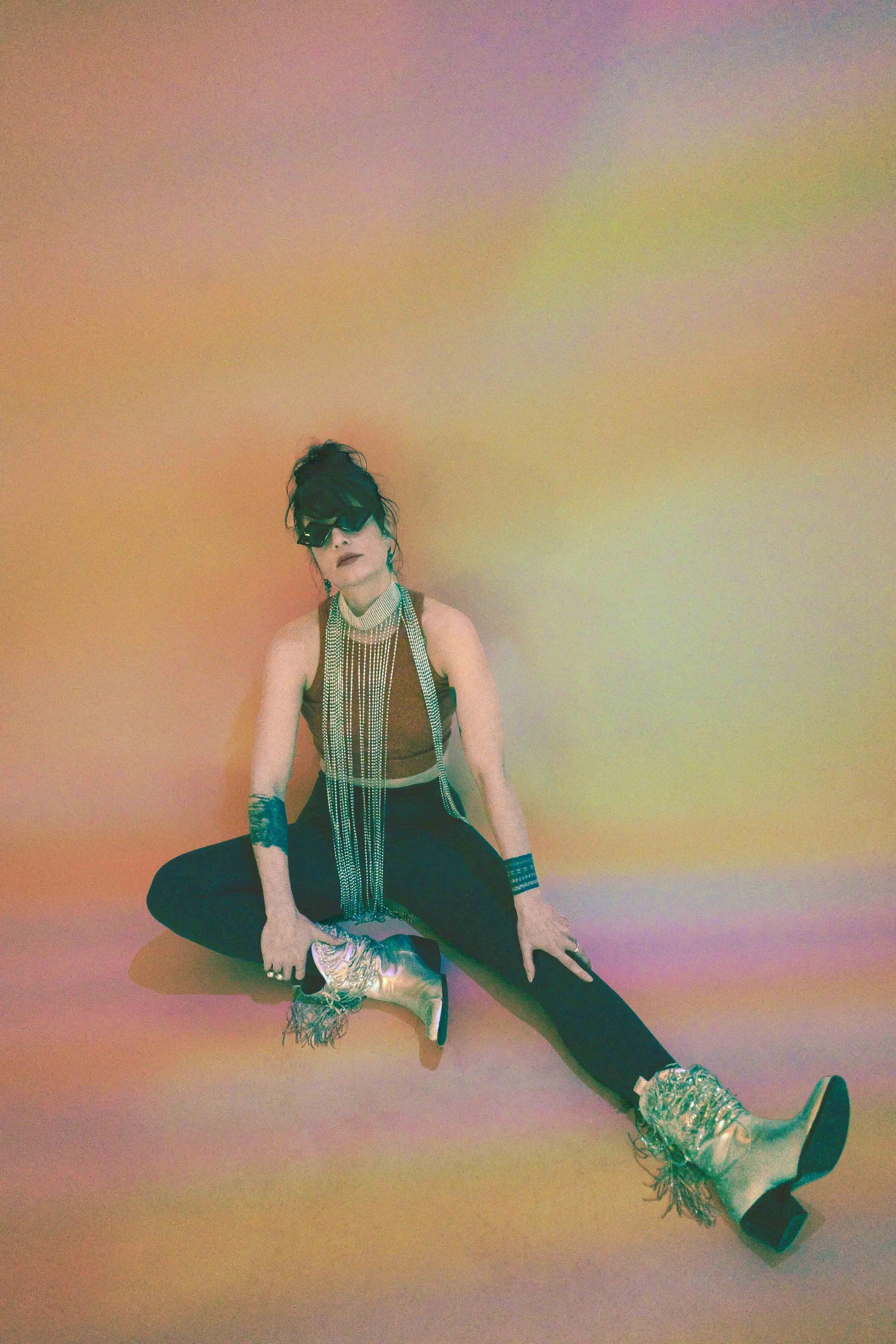 Woman sitting on the floor wearing dark sunglasses, metallic boots, and fashionable clothing with a colorful background.