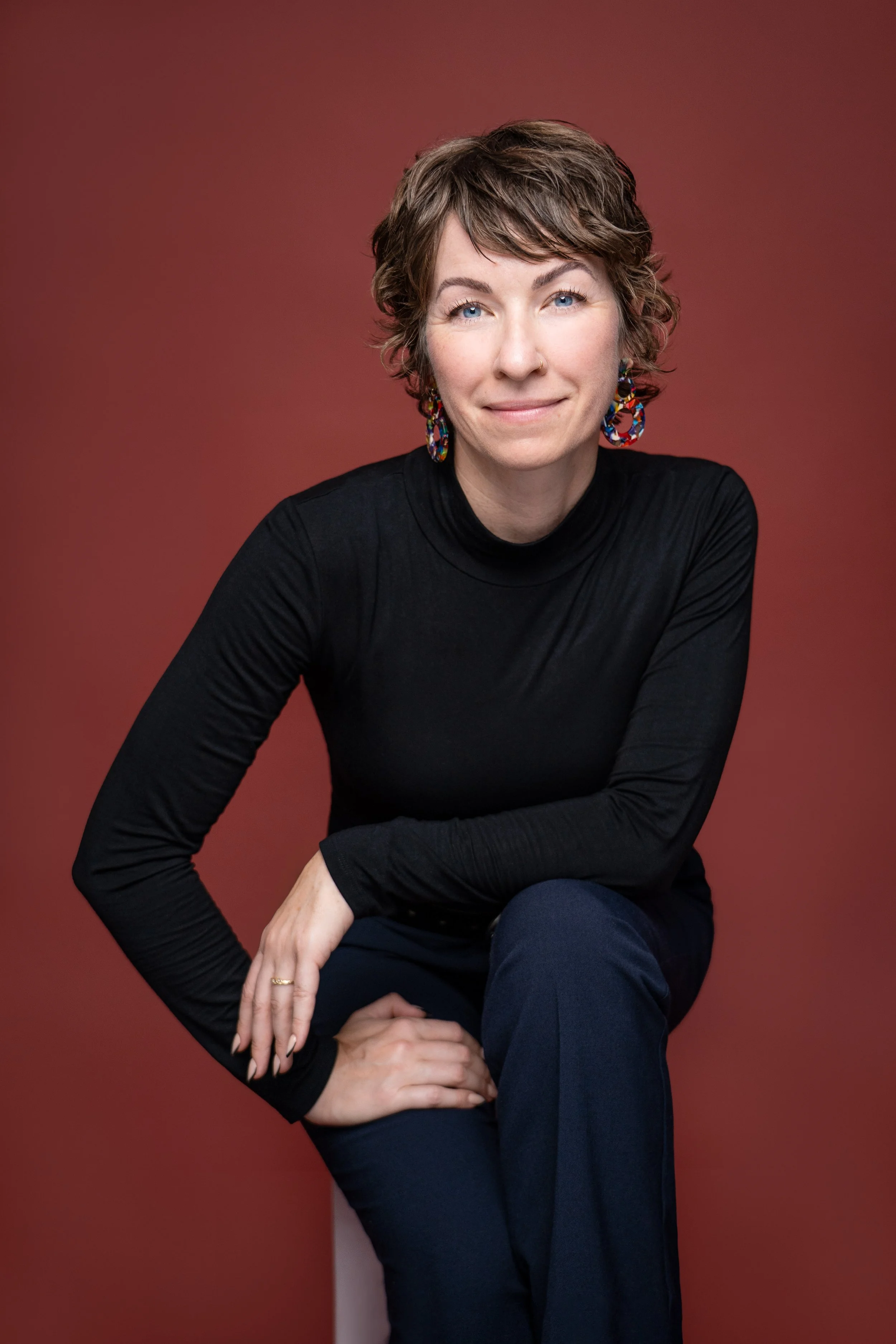 A woman with short curly brown hair, blue eyes, wearing a black turtleneck and colorful earrings, posing against a plain maroon background.