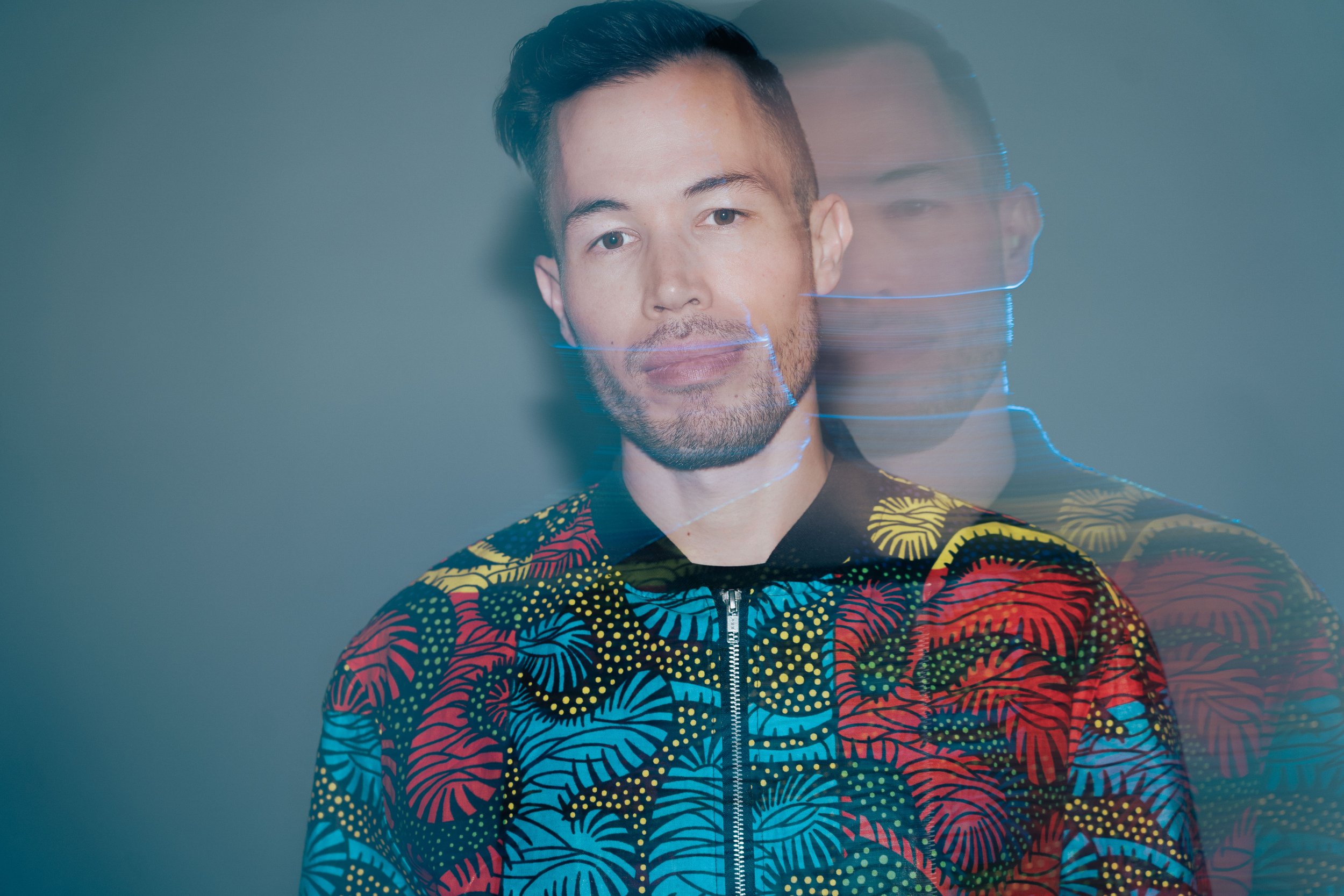 A young man with short dark hair and a beard posing against a plain background, wearing a colorful, patterned jacket with a zipper down the front.