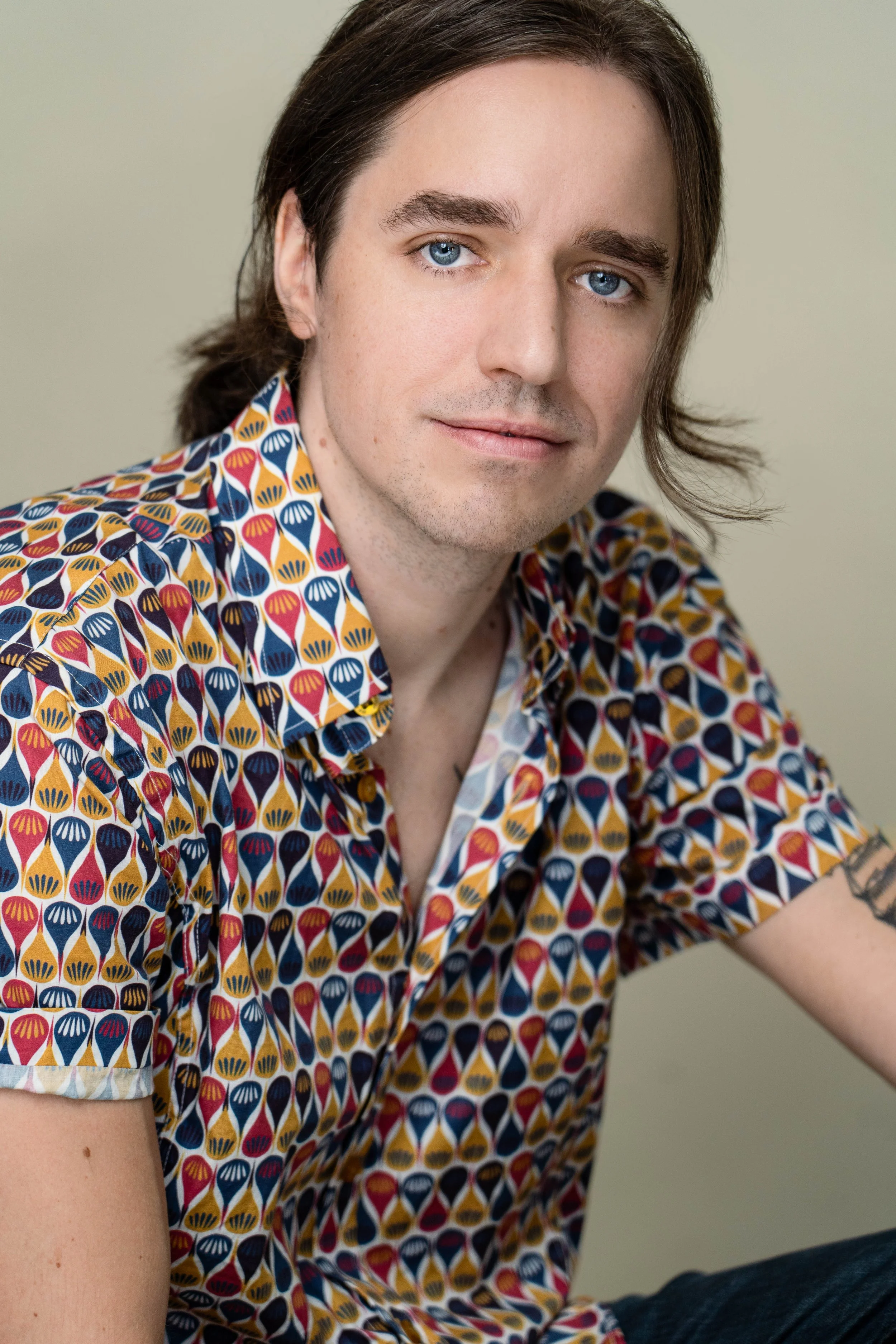 A young man with blue eyes and long brown hair, wearing a colorful patterned short-sleeve shirt, sitting against a plain neutral background.