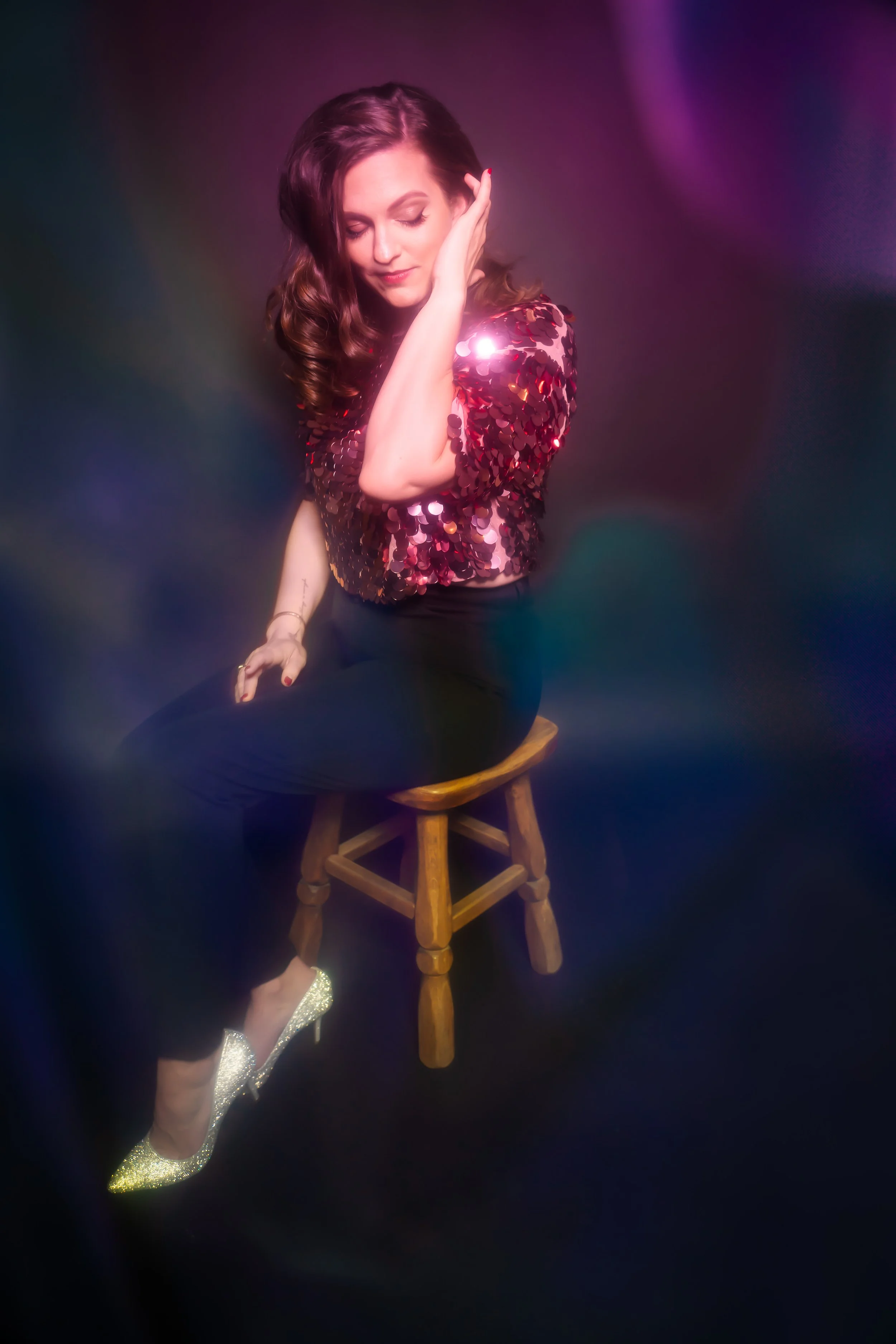 A woman with wavy brown hair sitting on a wooden stool studio photo in front of a dark background with colorful lights, wearing a shiny red sequin top, black pants, and glittery gold high heels.