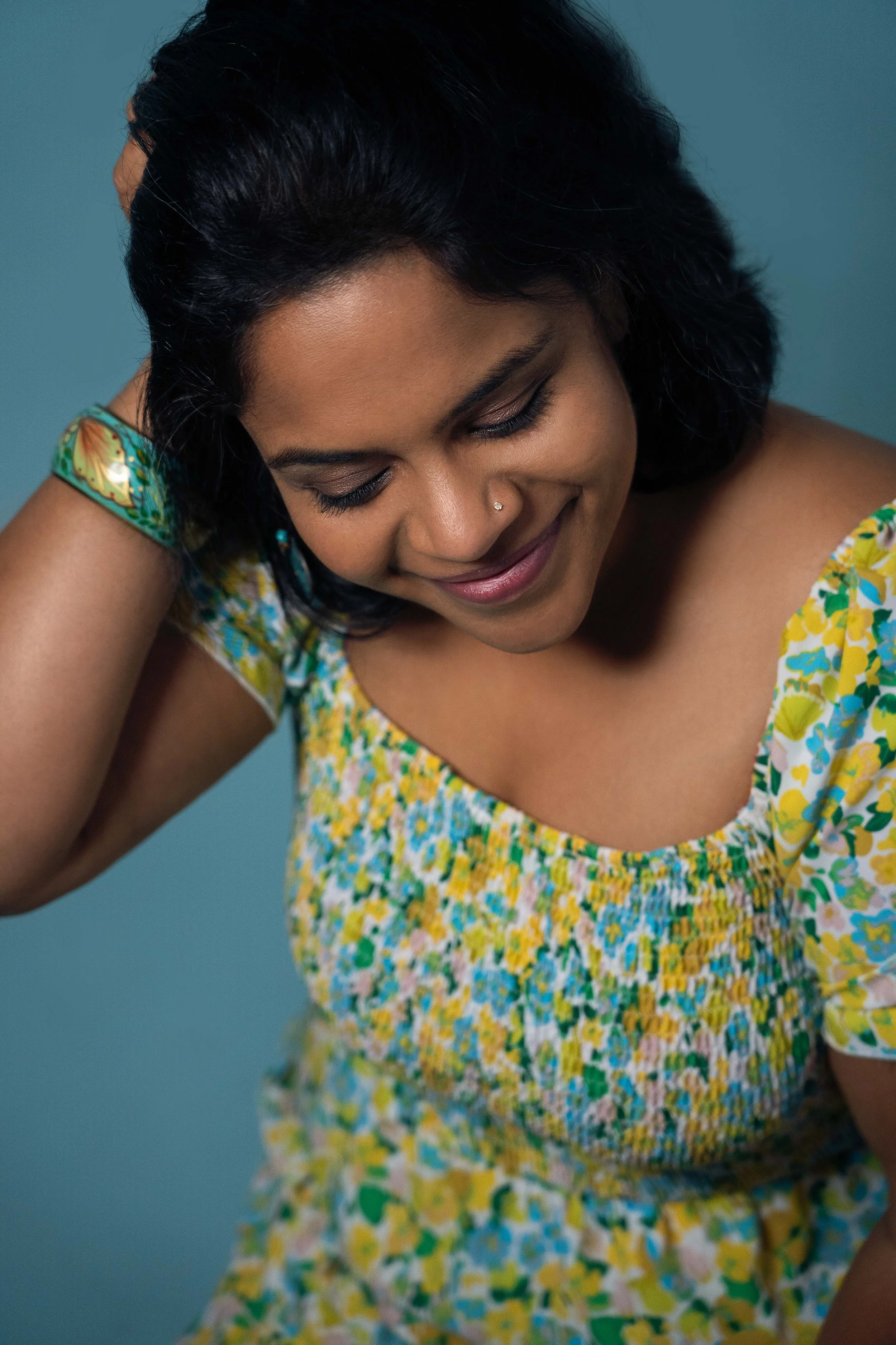 A woman with dark hair smiling, wearing a yellow floral dress, and a small nose piercing, with a blue background.