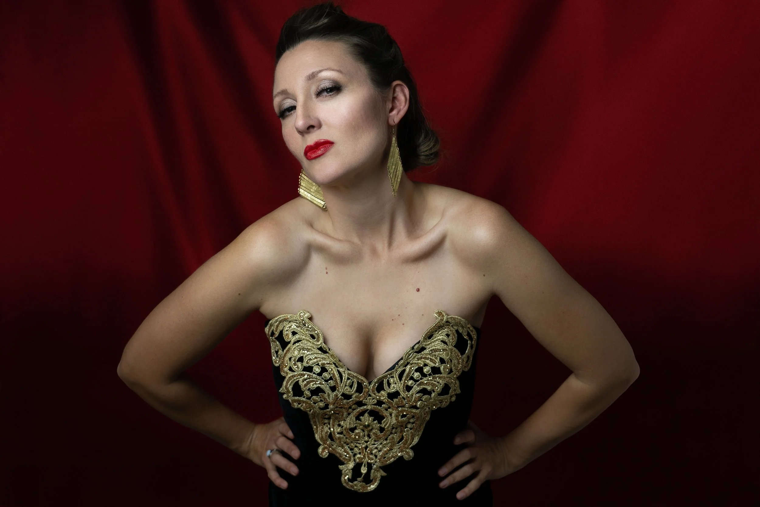 A woman with dark hair and red lipstick, wearing a black strapless dress with gold embroidery, stands with her hands on her hips against a red backdrop.