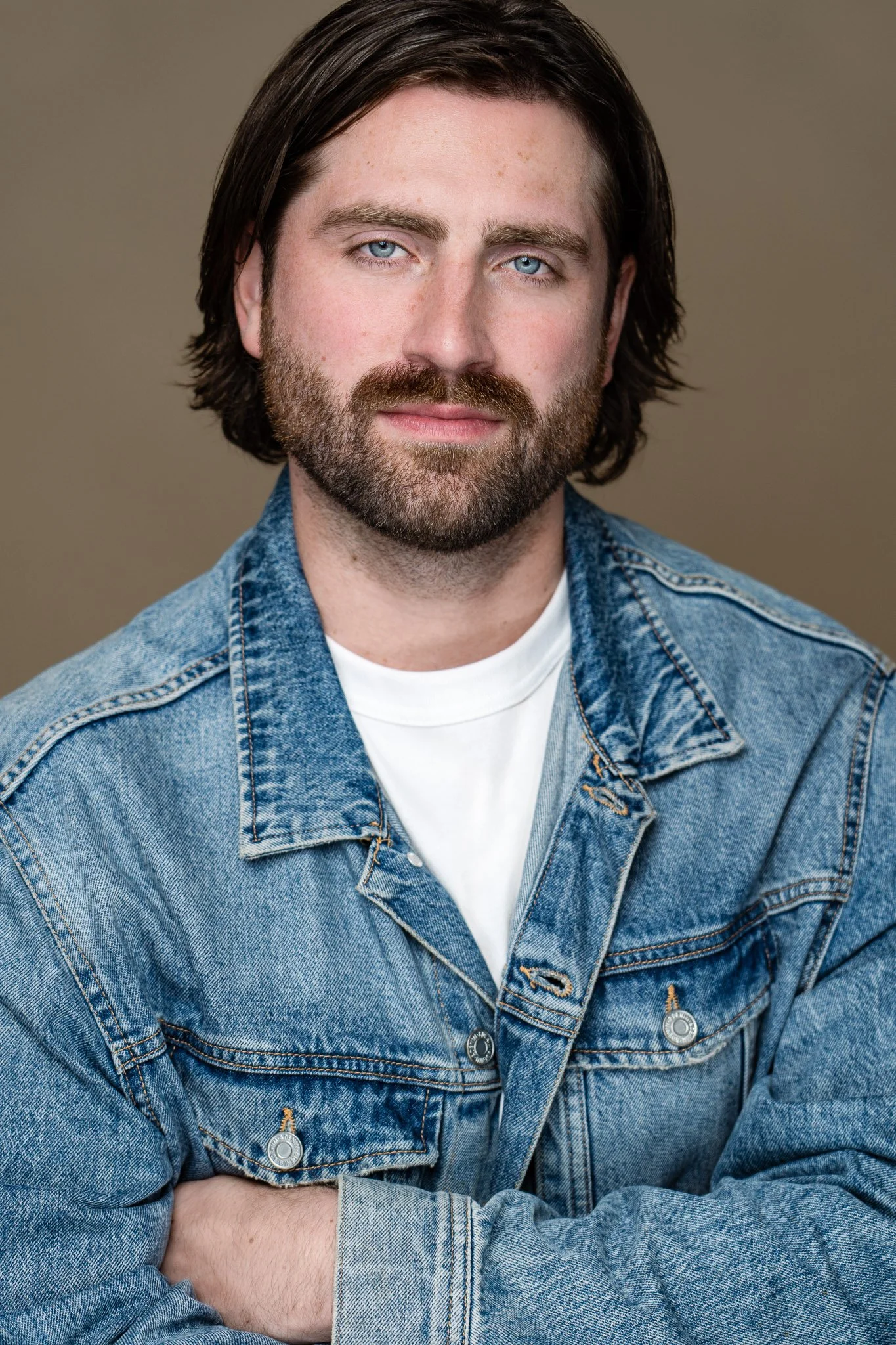 Portrait of a man with shoulder-length brown hair, blue eyes, and a beard, wearing a denim jacket over a white t-shirt, against a plain background.
