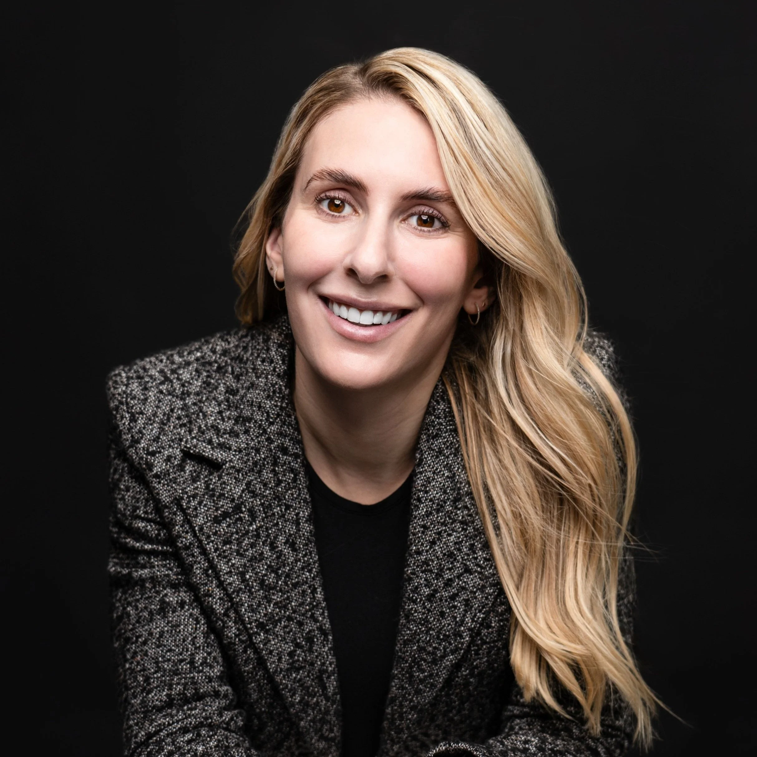 Professional woman with long blonde wavy hair smiling in front of black background.