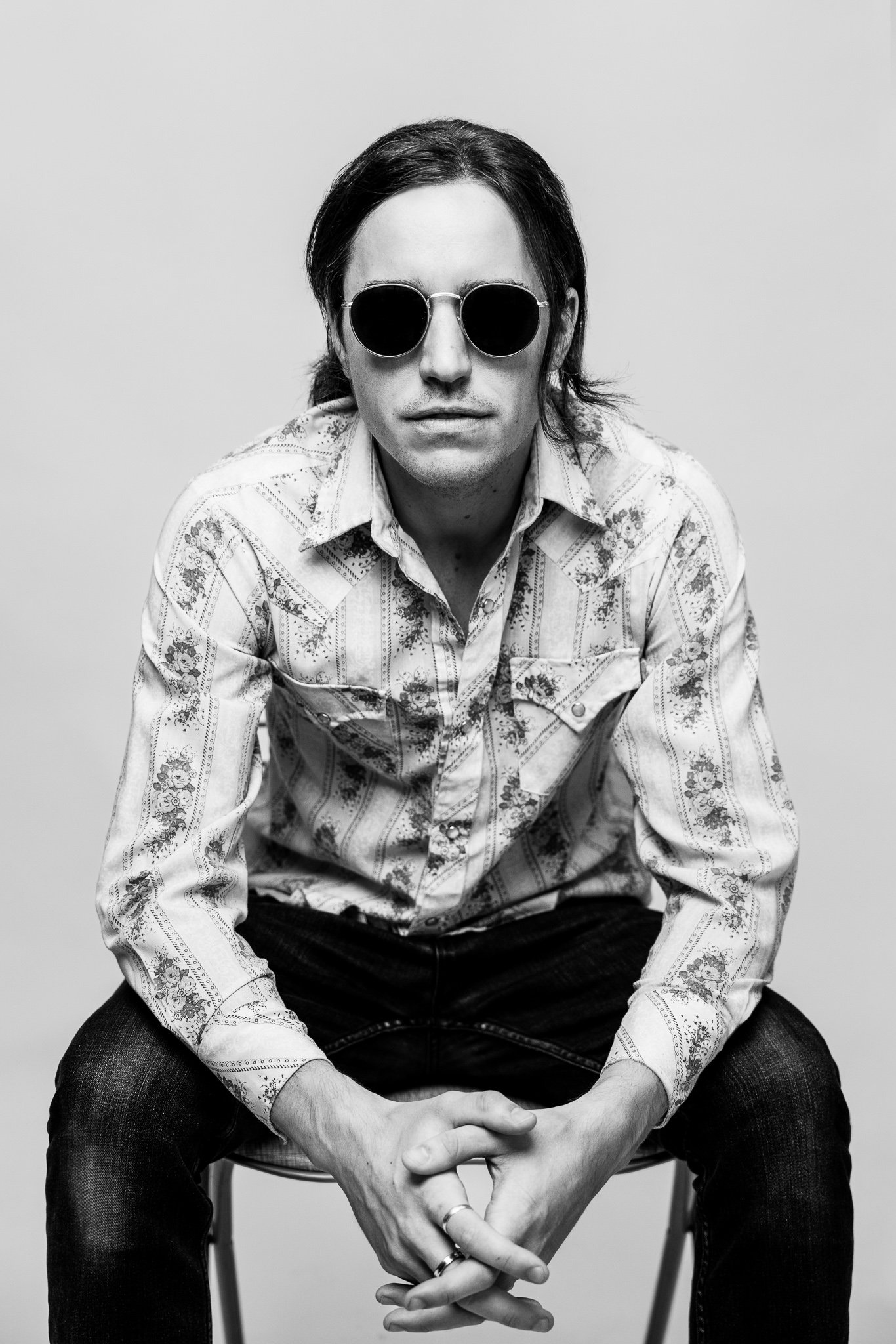 Black and white portrait of a man with shoulder-length hair wearing round dark sunglasses, a patterned button-up shirt, and dark jeans, seated and looking directly at the camera.