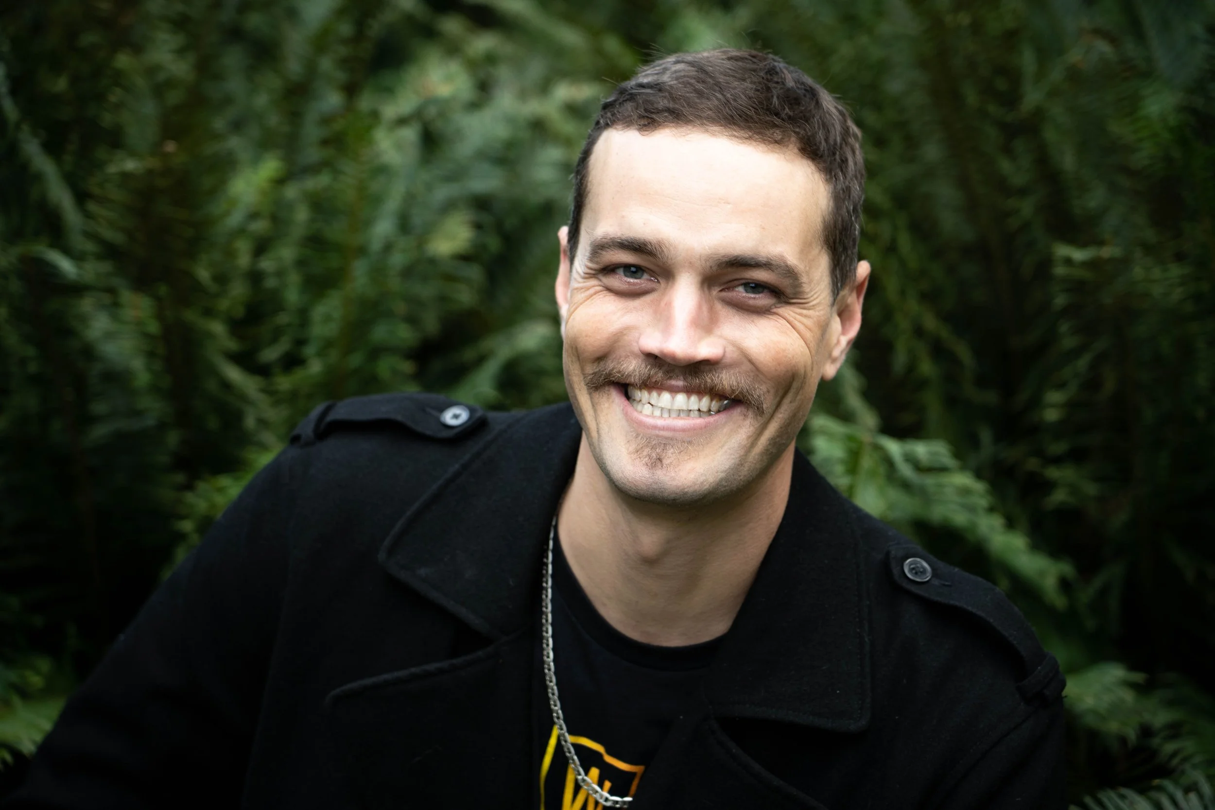 A smiling man with short brown hair, a goatee, wearing a black jacket and necklace, outdoors with green foliage in the background.