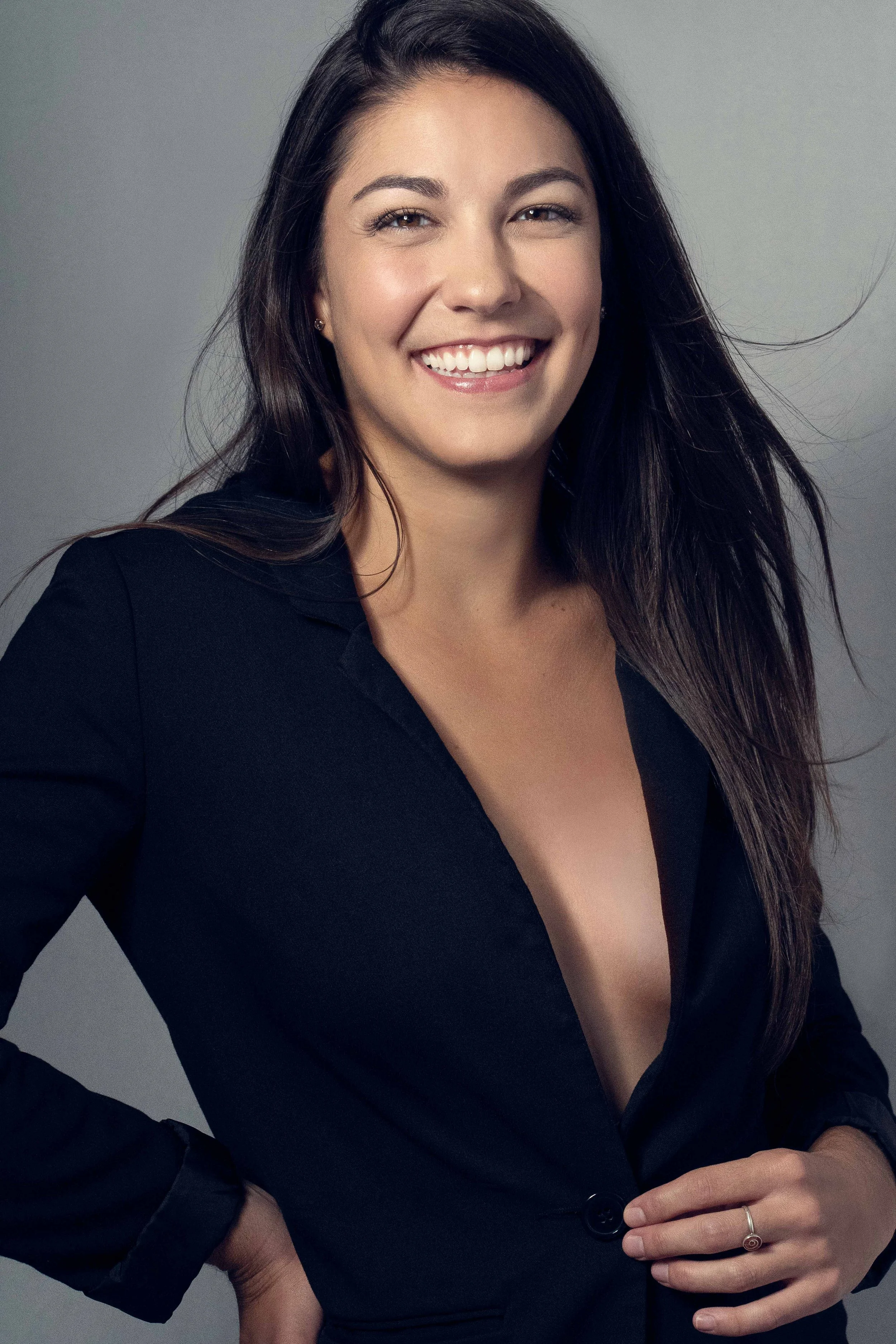 A woman with long dark hair, wearing a black blazer with a deep neckline, smiling and posing with one hand on her hip against a neutral background.