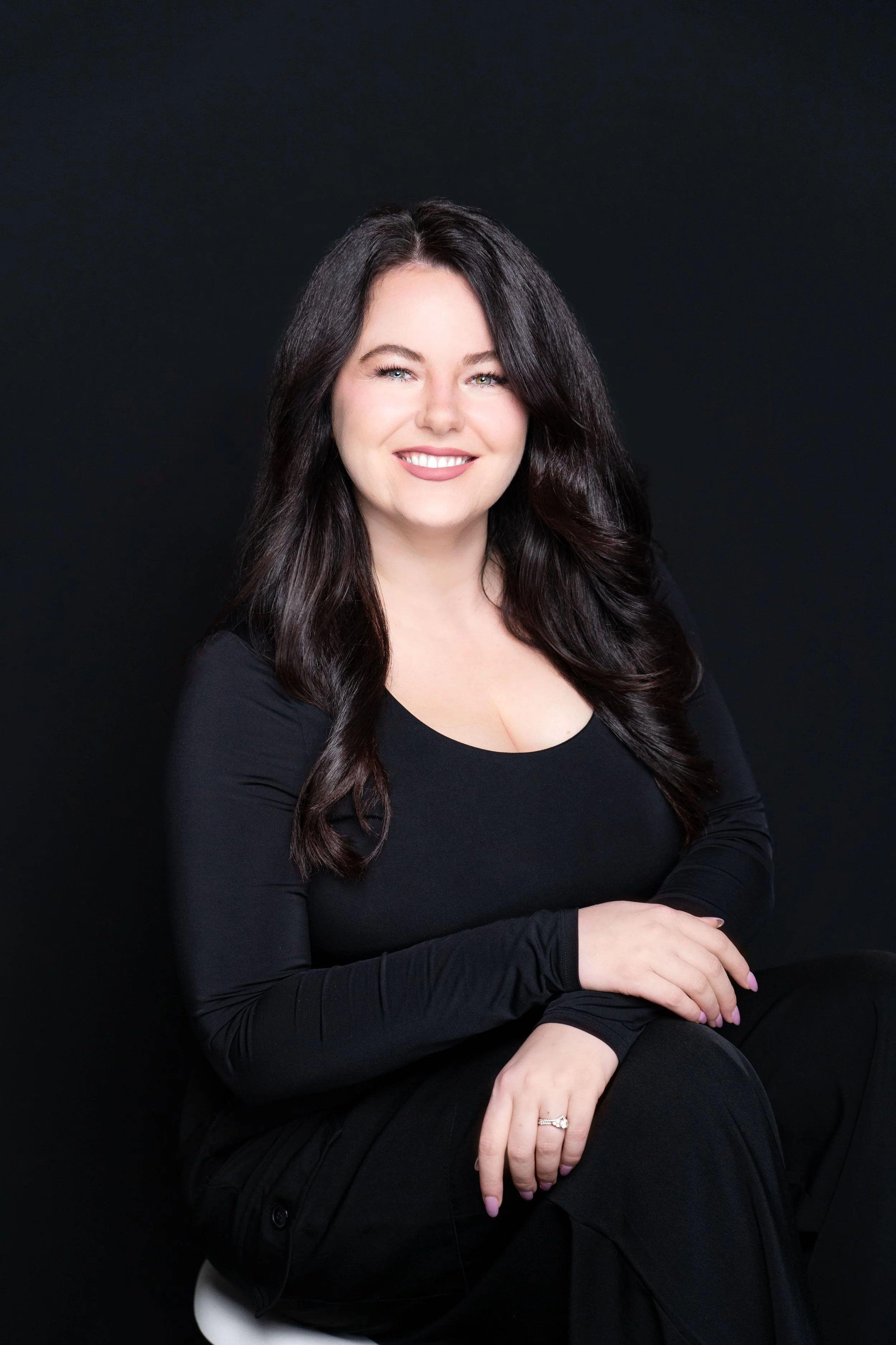 Portrait of a smiling woman with long dark hair, wearing a black top and sitting against a plain black background.