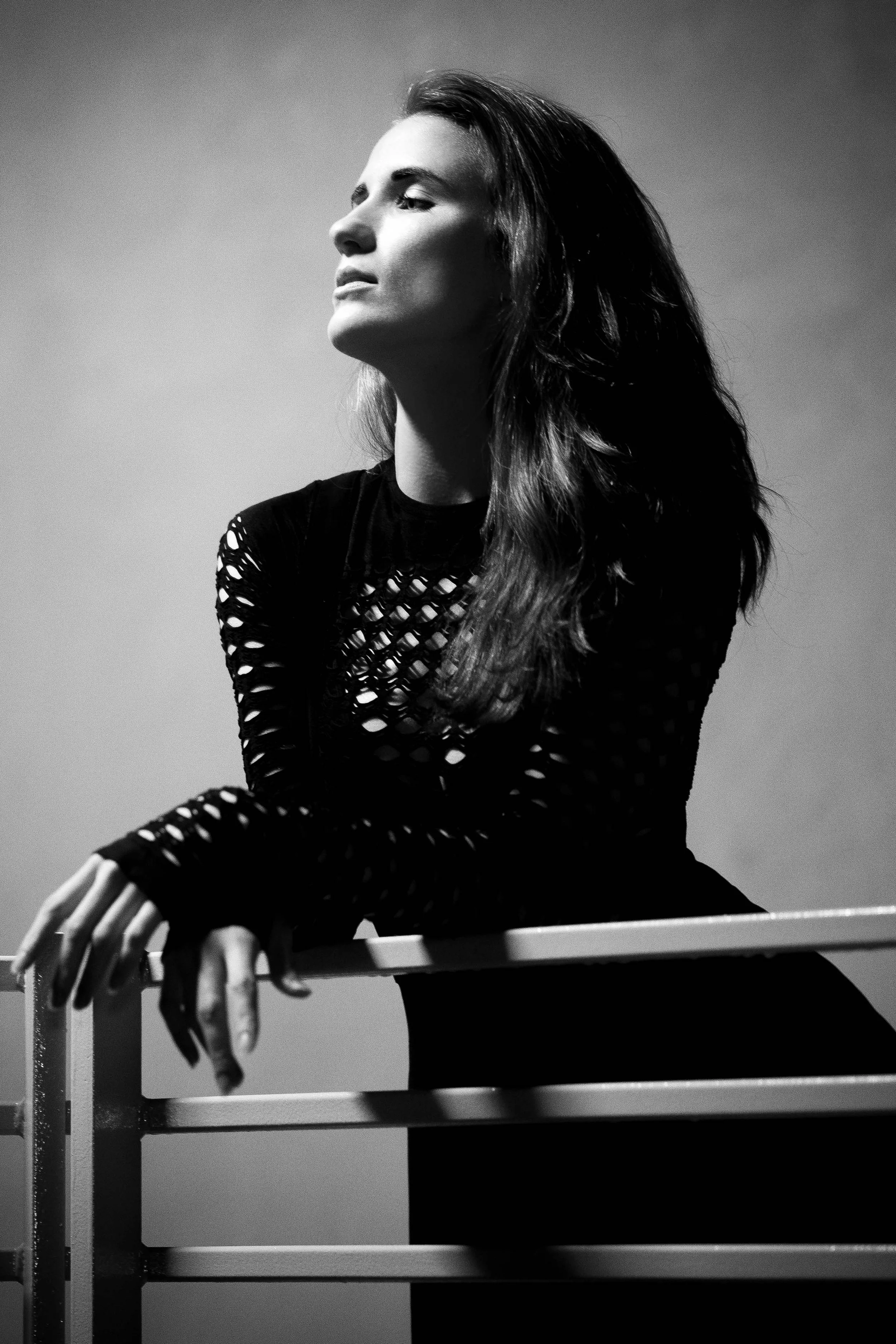 Black and white portrait of a woman with long hair, looking to the side, leaning on a railing, wearing a mesh top.