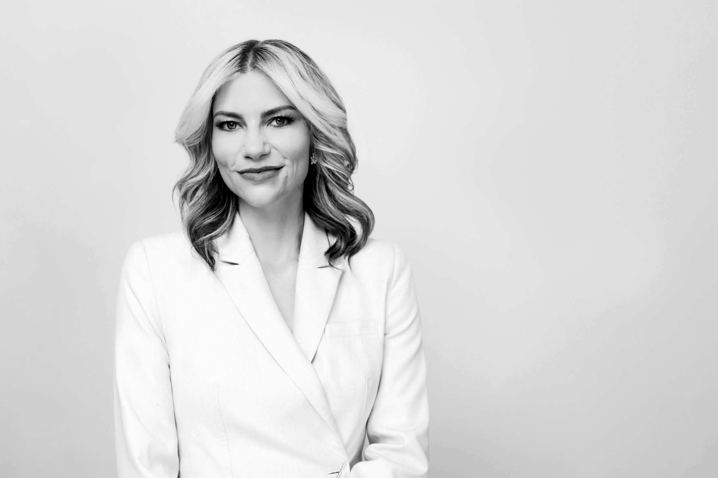Black and white portrait of a woman with shoulder-length wavy hair, wearing a white blazer, smiling softly, against a plain light background.