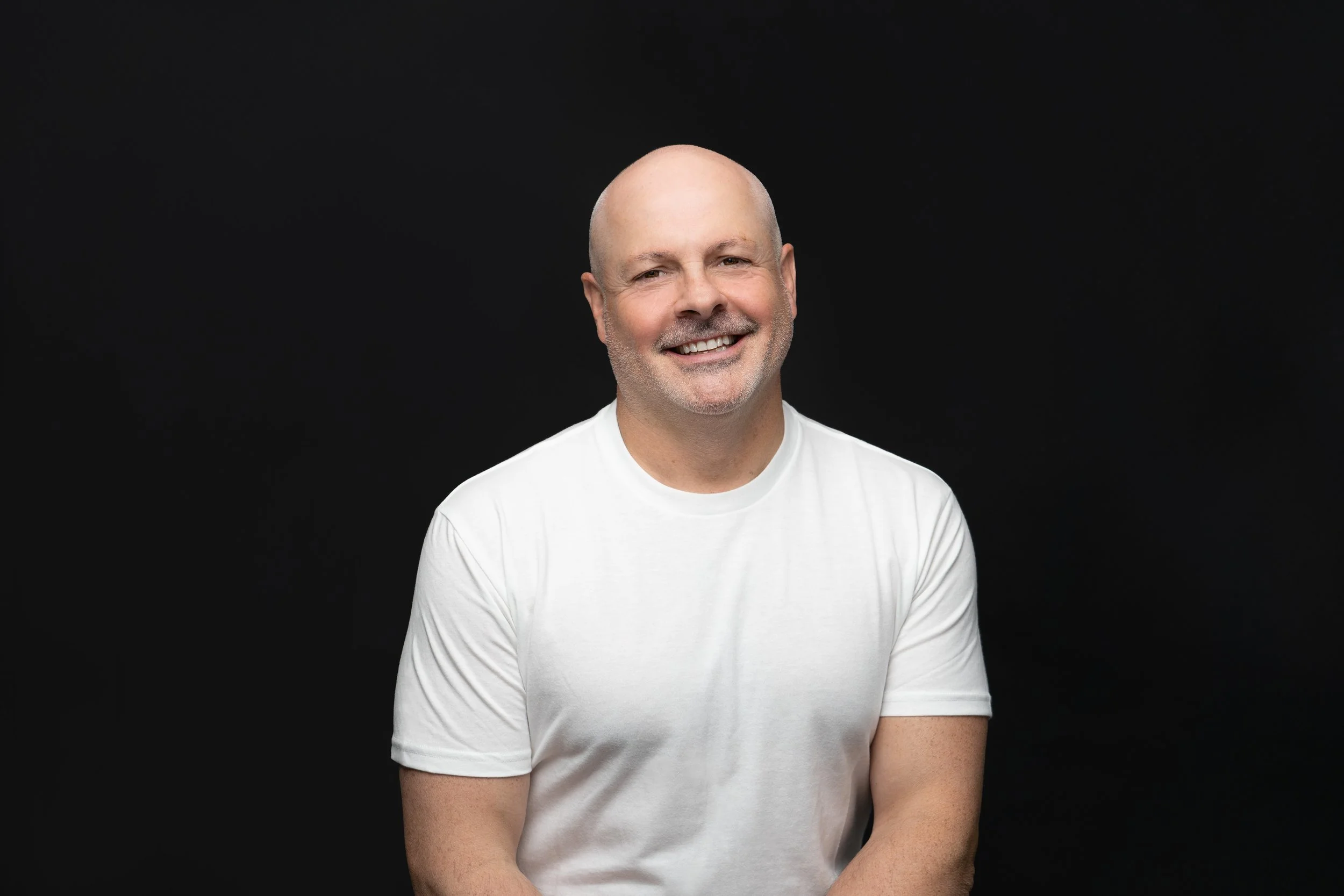 A smiling bald man with a beard wearing a white t-shirt against a black background.