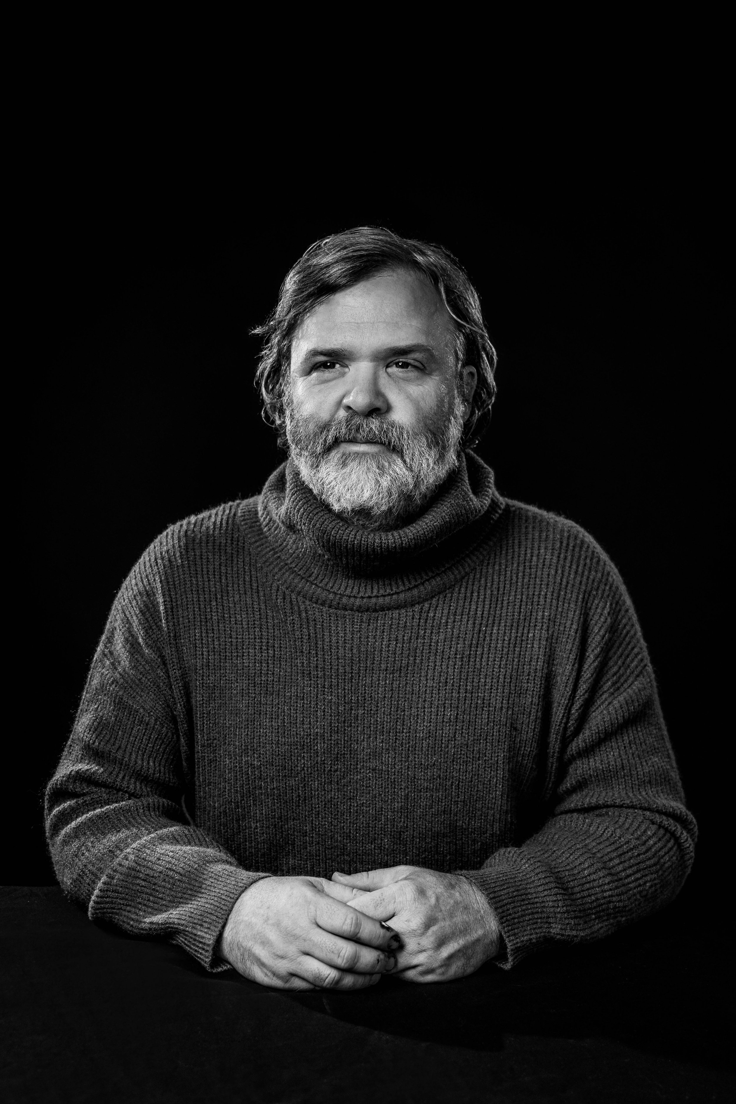 Black and white portrait of a middle-aged man with a beard and wavy hair, wearing a turtleneck sweater, sitting at a dark table against a black background.