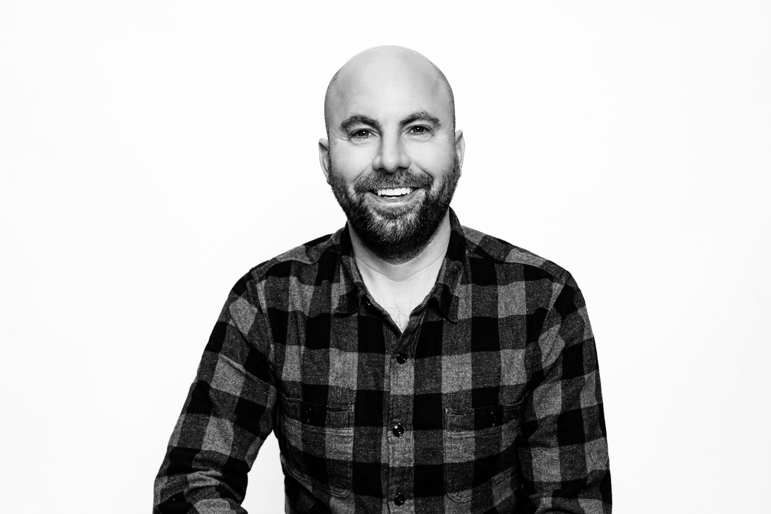 A smiling man with a beard and bald head, wearing a black and gray plaid shirt, posing against a plain white background.
