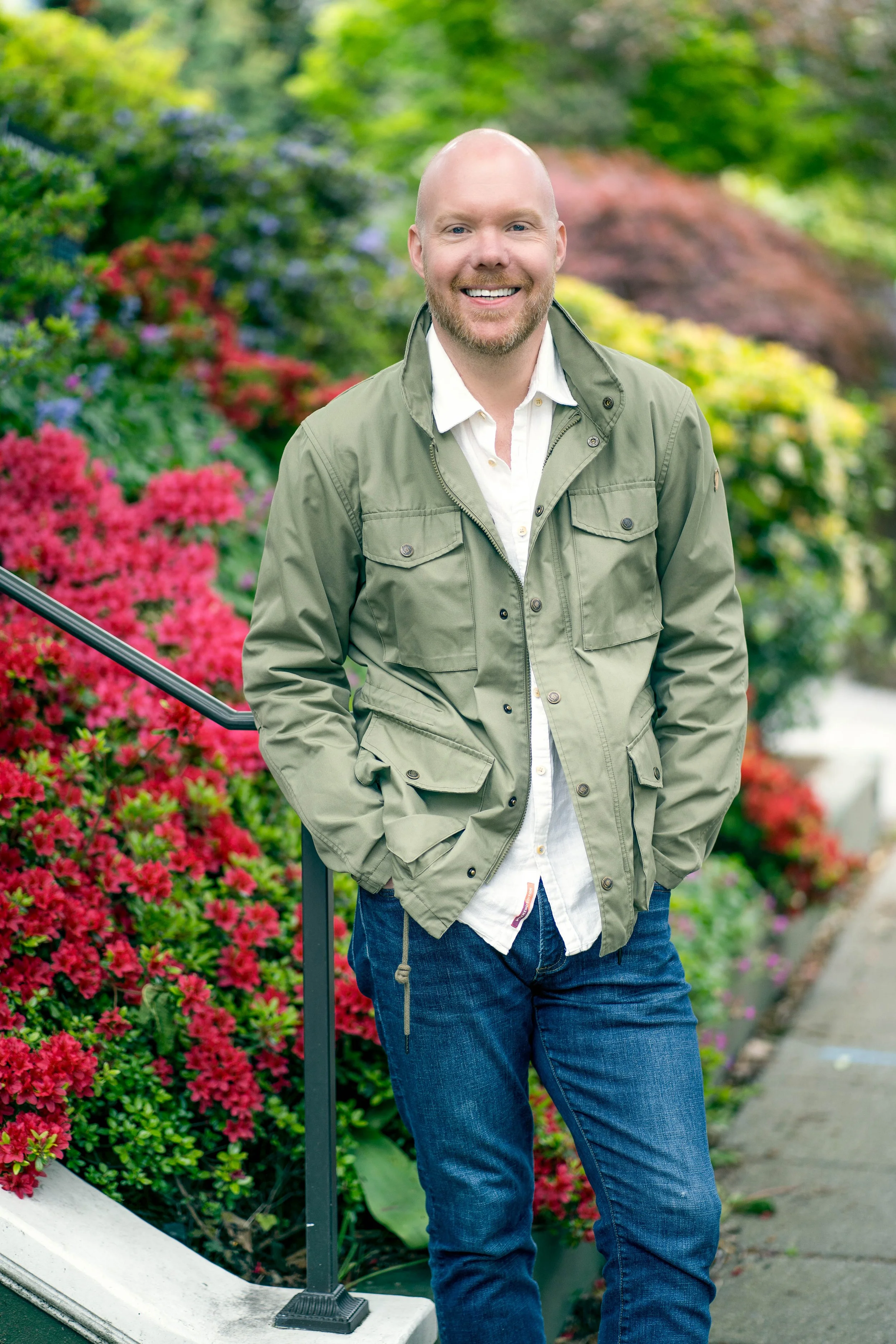 A man with a bald head and beard smiling outdoors, standing in front of colorful blooming flowers and greenery.