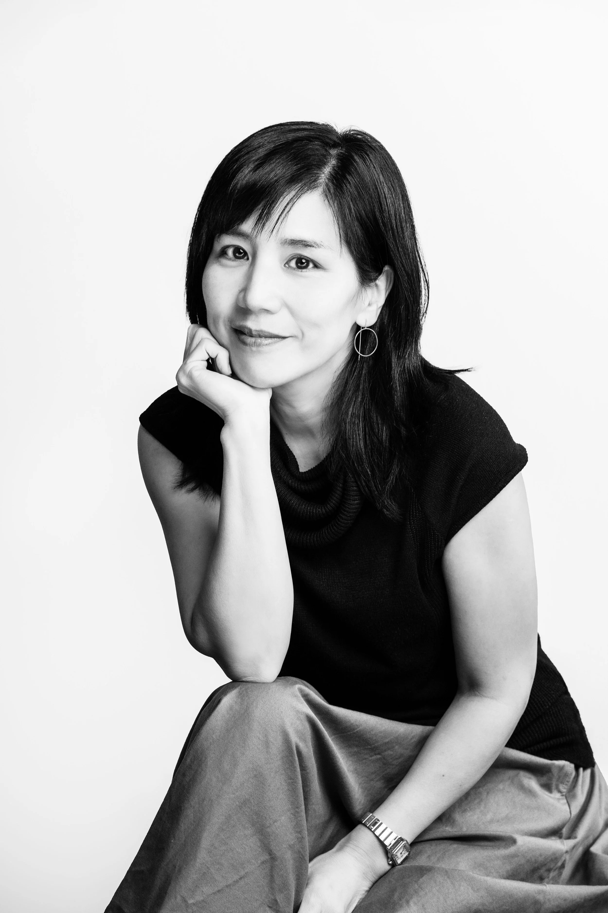 A black-and-white portrait of an Asian woman smiling, with chin resting on her hand, wearing a black sleeveless top, large hoop earrings, and a wristwatch.