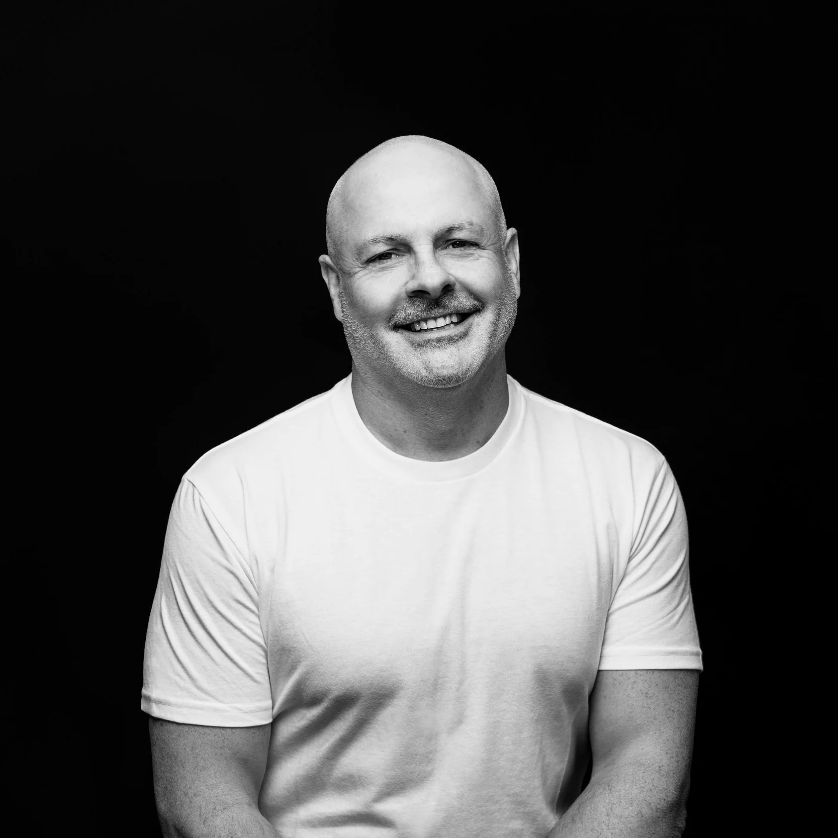 Black and white portrait of a smiling man with a shaved head and a beard, wearing a white t-shirt against a dark background.