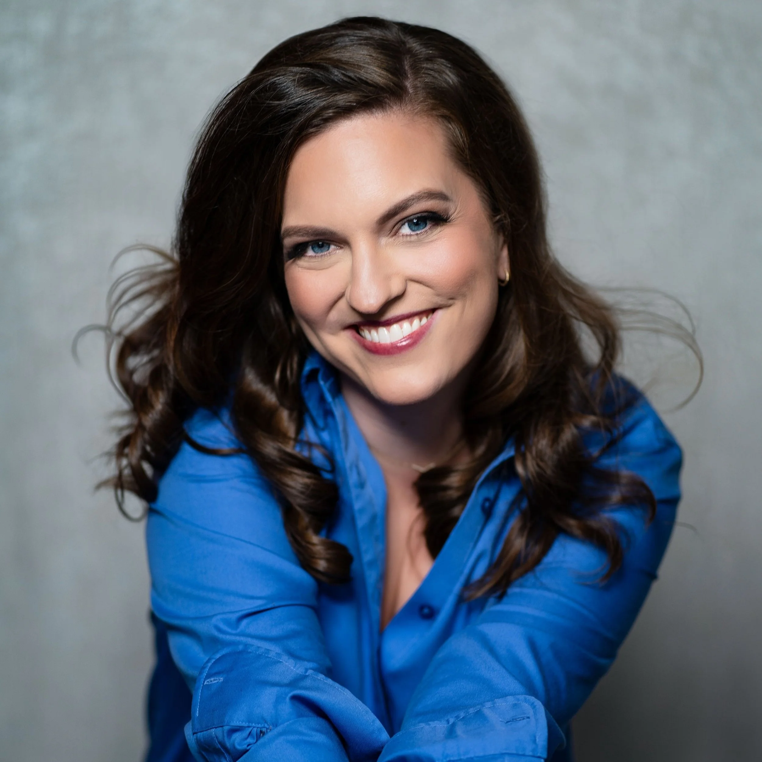 Close-up of a smiling woman with long, wavy brown hair, wearing a blue shirt, against a neutral background.