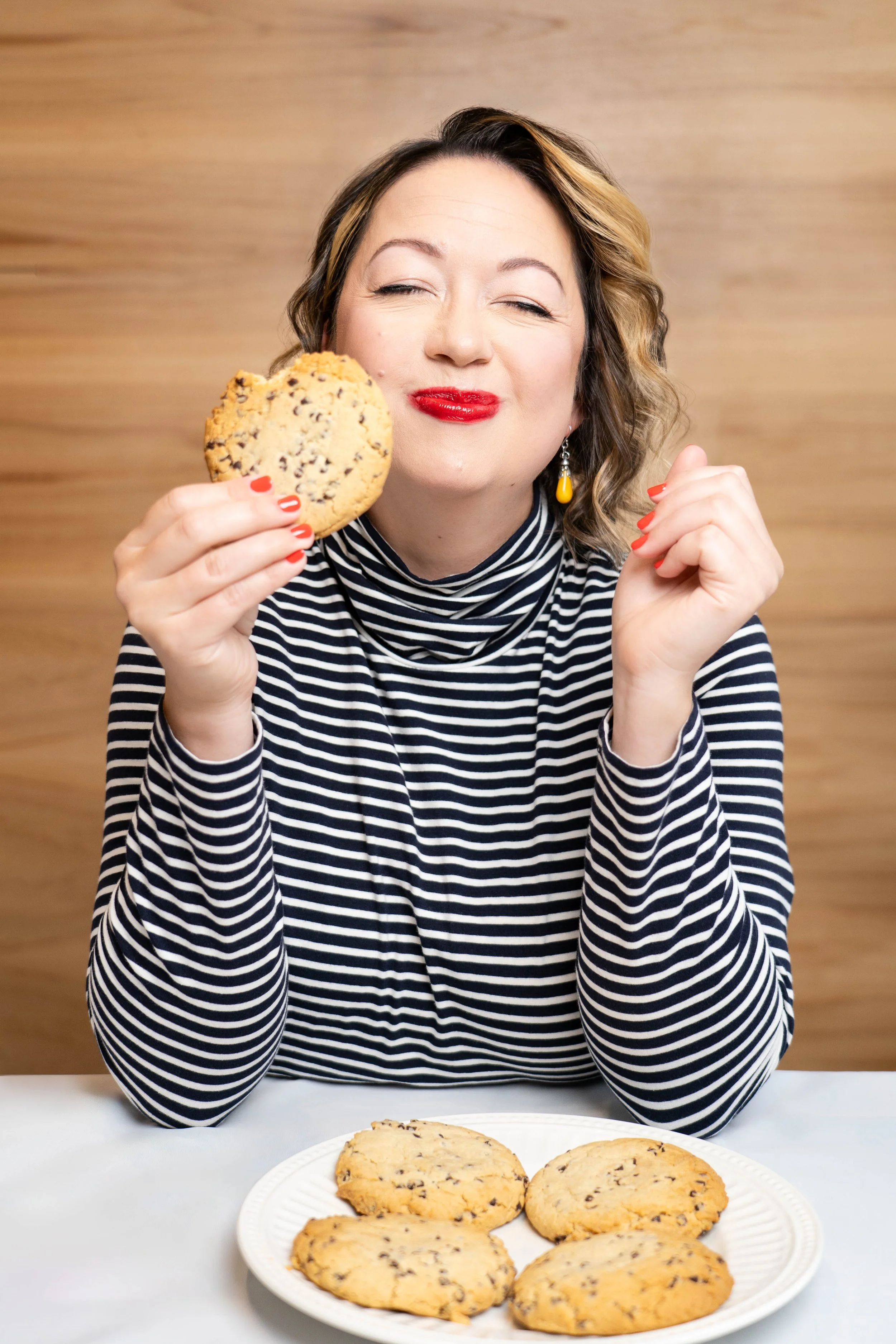 A woman with wavy brown hair and red lipstick sitting at a table holding a chocolate chip cookie up to her face. There is a plate with more cookies in front of her, and she is wearing a black and white striped long-sleeve shirt. The background is a l
