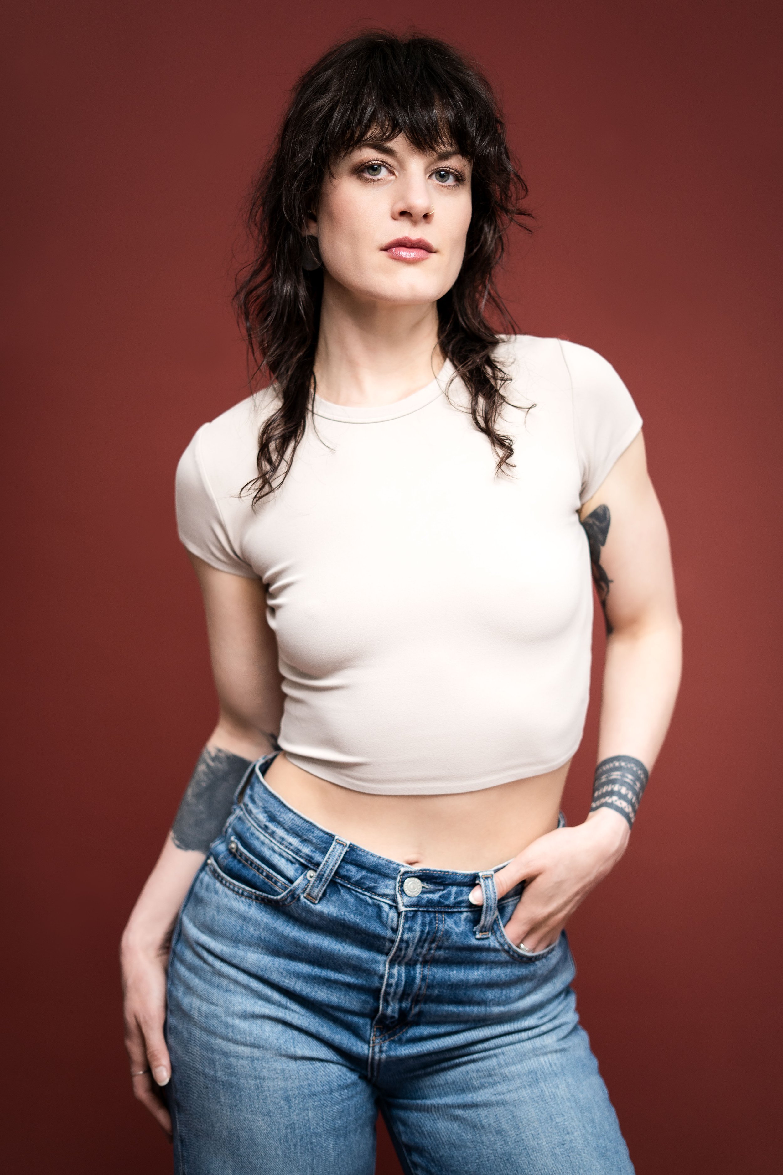 A woman with dark, wavy hair and tattoos on her arms poses against a plain reddish-brown background. She is wearing a light-colored, cropped t-shirt and high-waisted blue jeans, with one hand resting in her pocket.