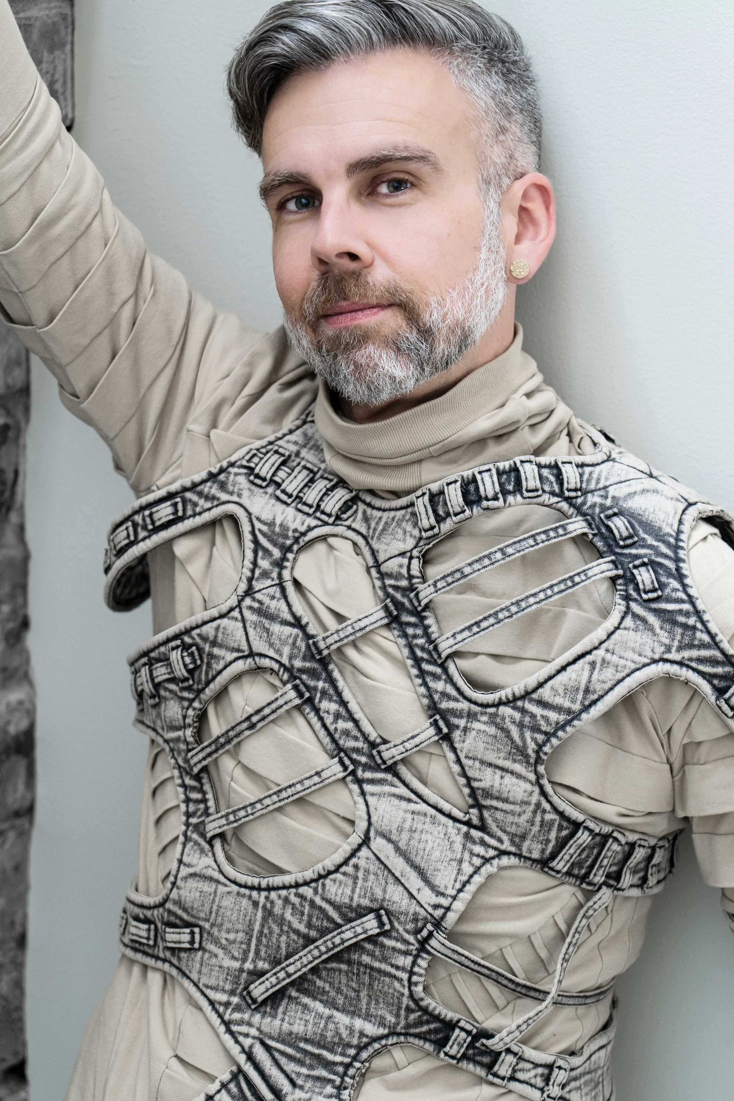 A man with gray hair and beard, wearing a beige turtleneck and a unique, layered jacket, standing against a white wall.