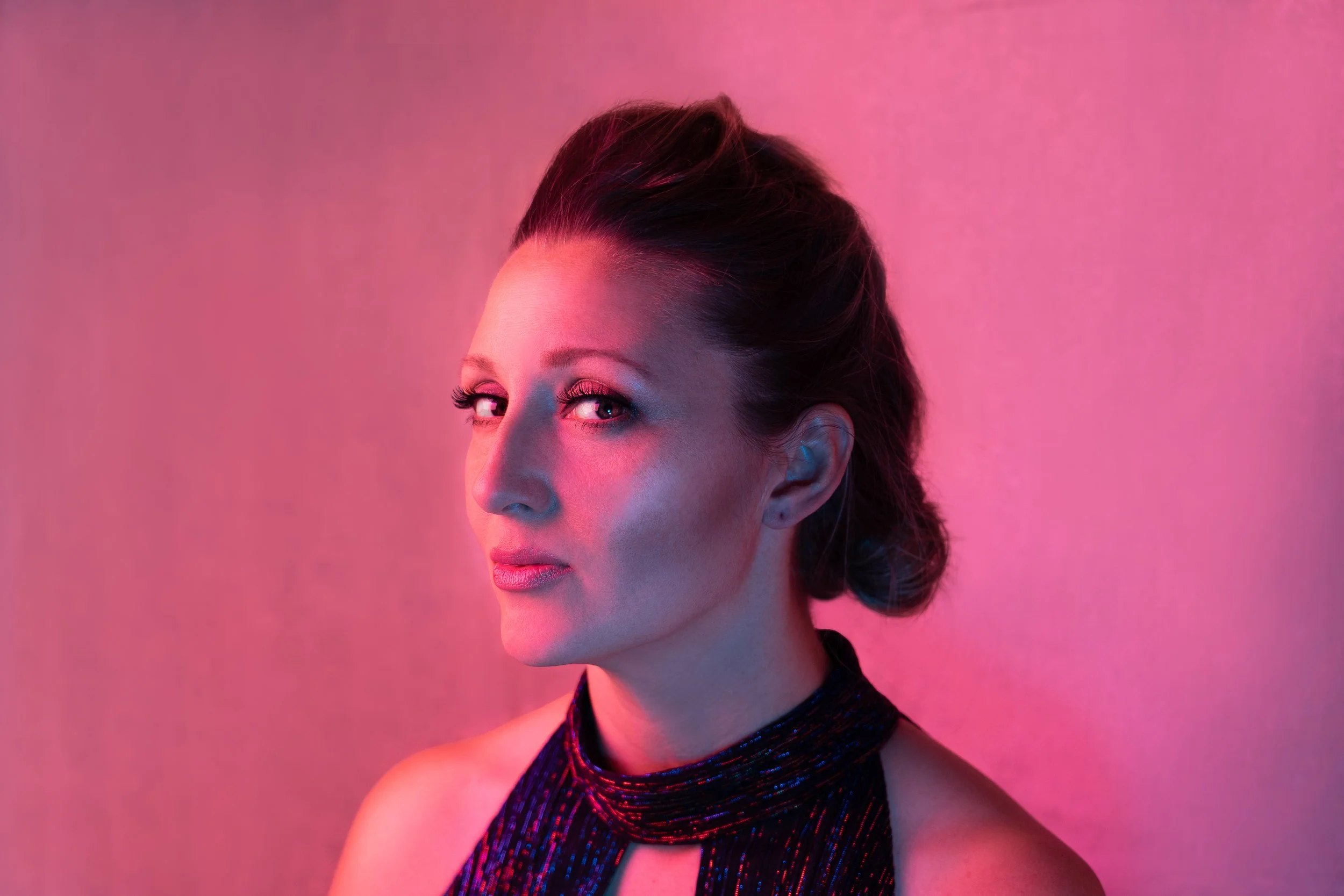 A woman with dark hair styled in a loose bun, looking over her shoulder with a confident expression, illuminated by pink and purple lighting.