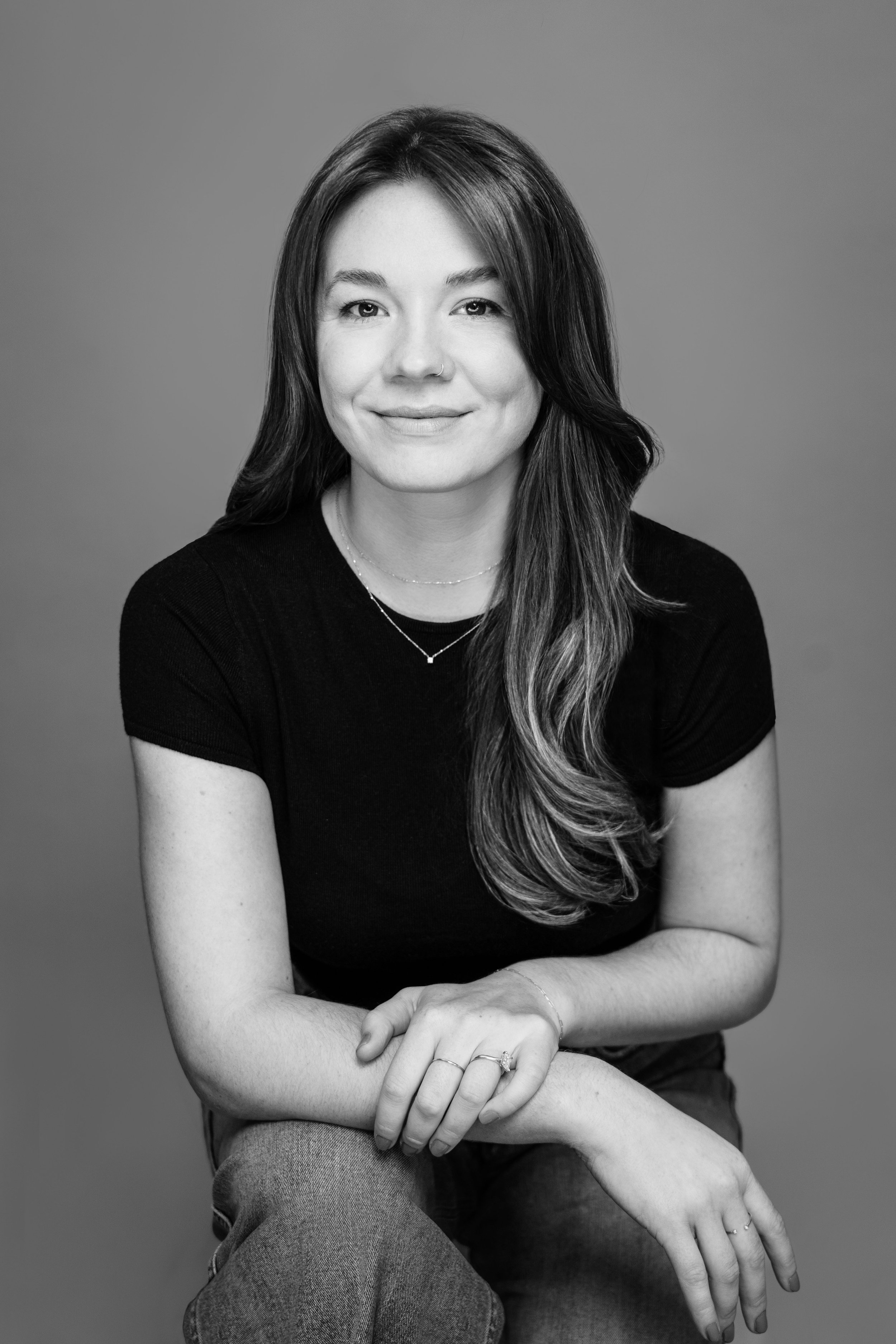 Black and white portrait of a woman with long hair, smiling, wearing a black top and jeans, sitting with her arms crossed.