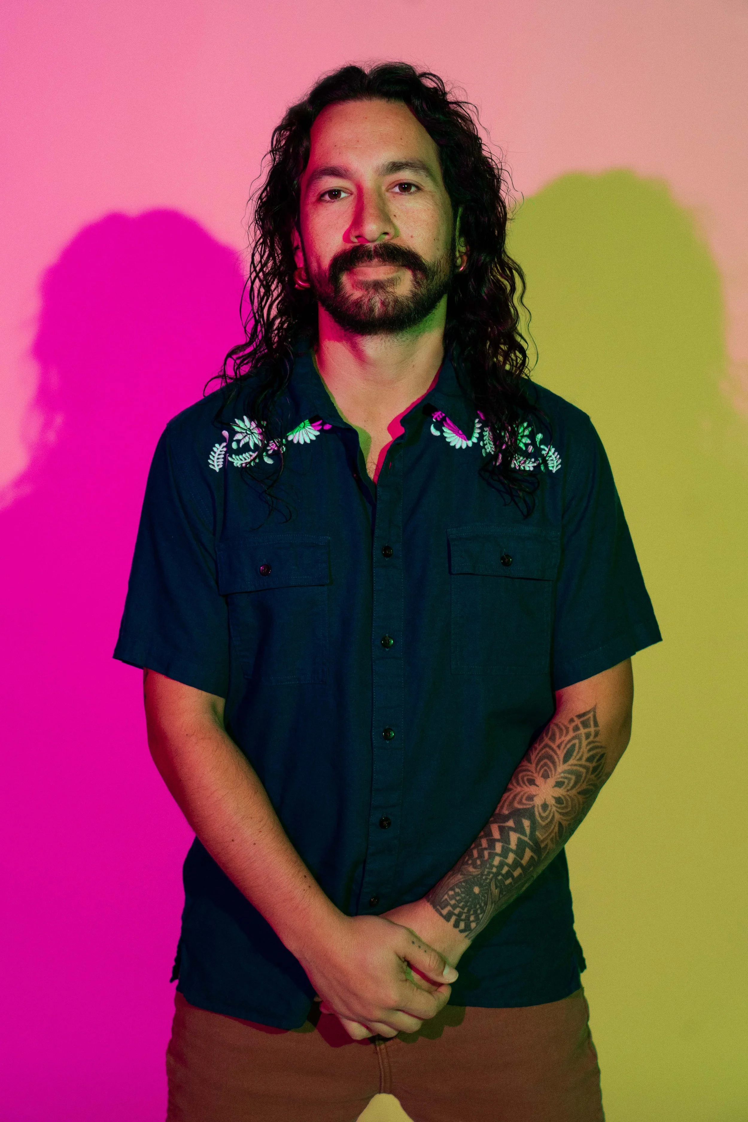 A man with long curly hair, beard, and tattoos on his left arm, standing against a wall with pink and yellow lighting shadows, wearing a dark short-sleeved shirt with floral embroidery.