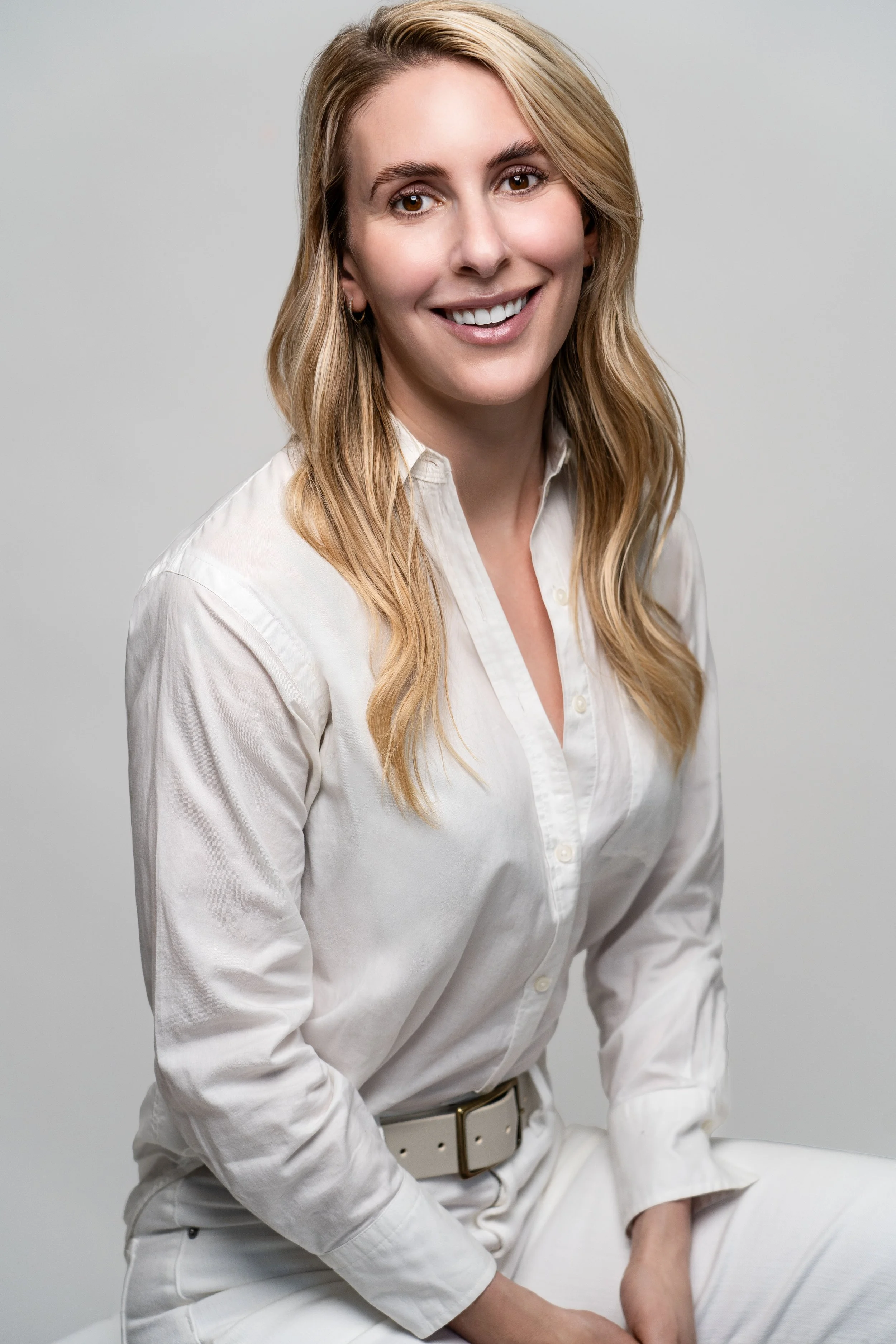 A smiling woman with blonde hair wearing a white button-up shirt and white pants against a light gray background.