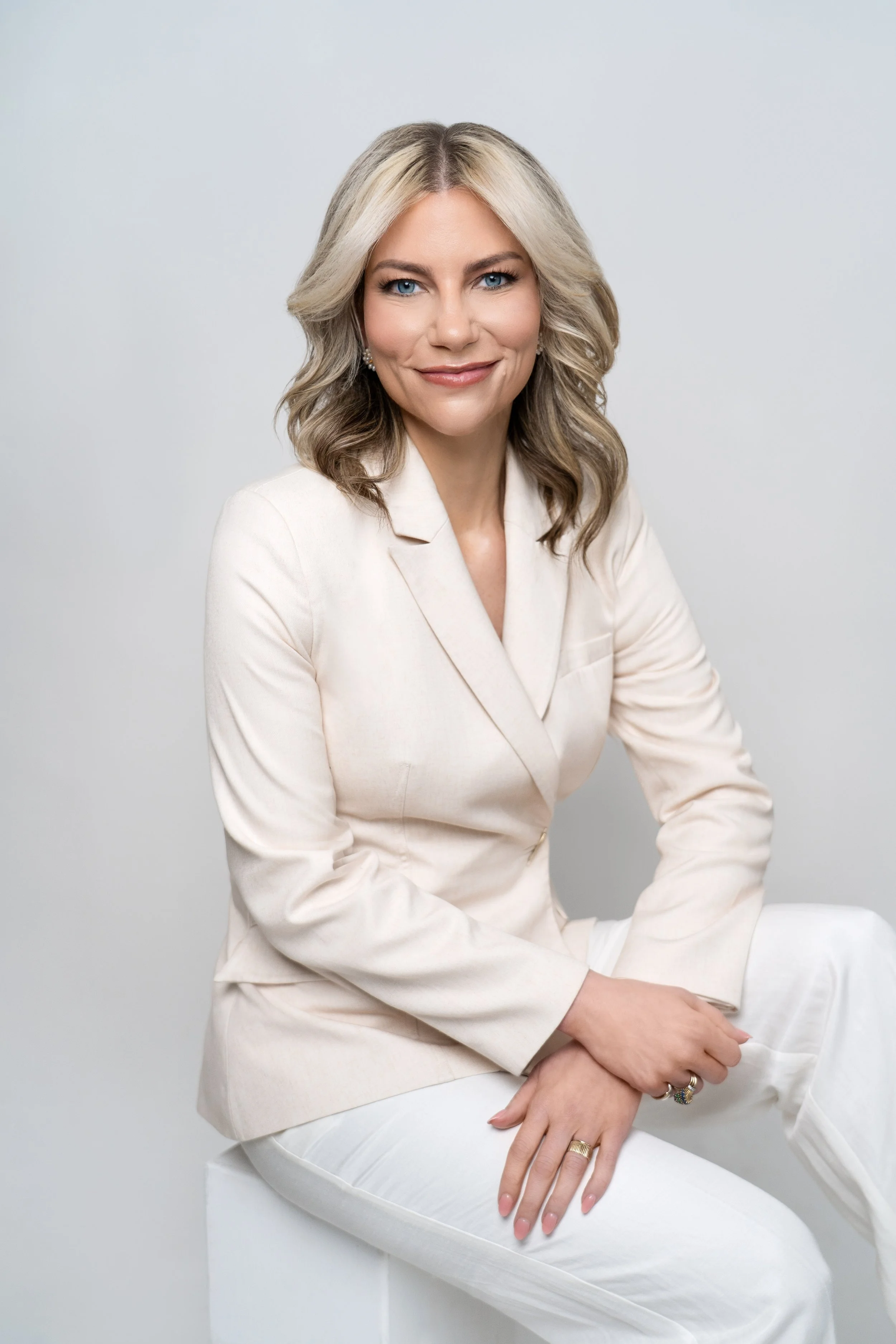 A woman with blonde, wavy hair smiling, wearing a cream-colored blazer and white pants, sitting on a white block against a light gray background.