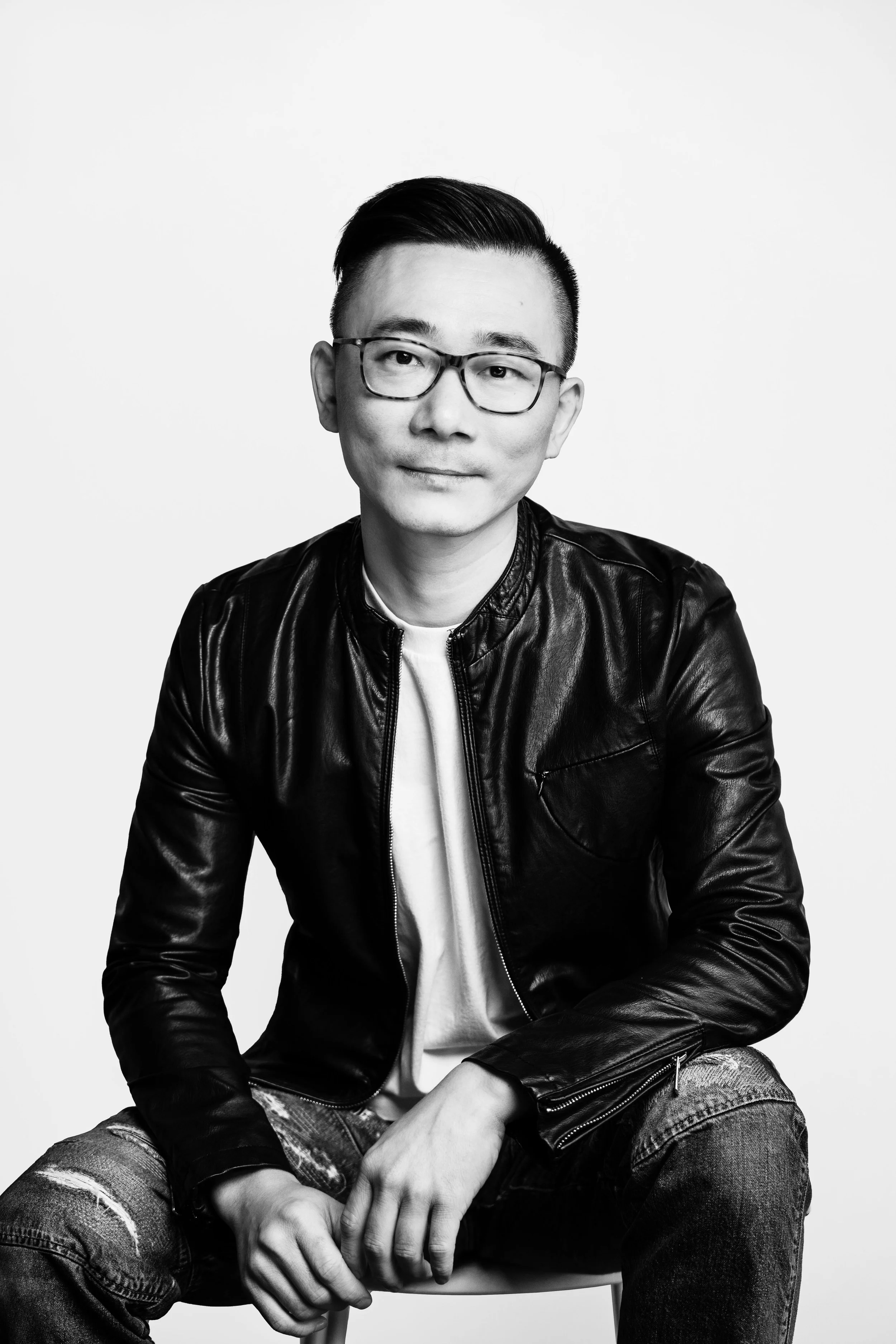 Black and white portrait of a young man with glasses, wearing a leather jacket and jeans, sitting on a chair against a plain white background.