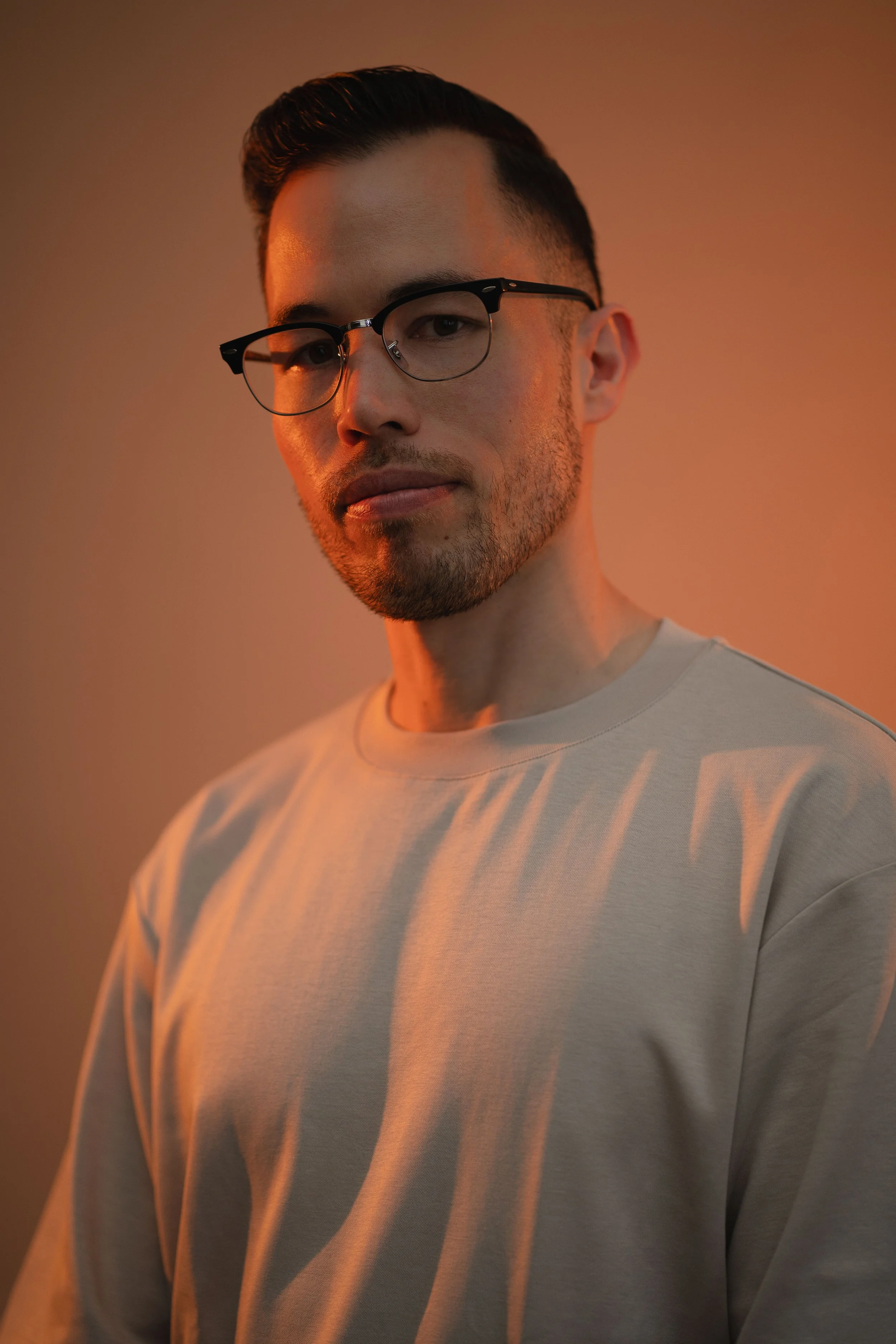 Portrait of a young man wearing glasses and a beige sweatshirt, with warm lighting highlighting his face against a plain background.