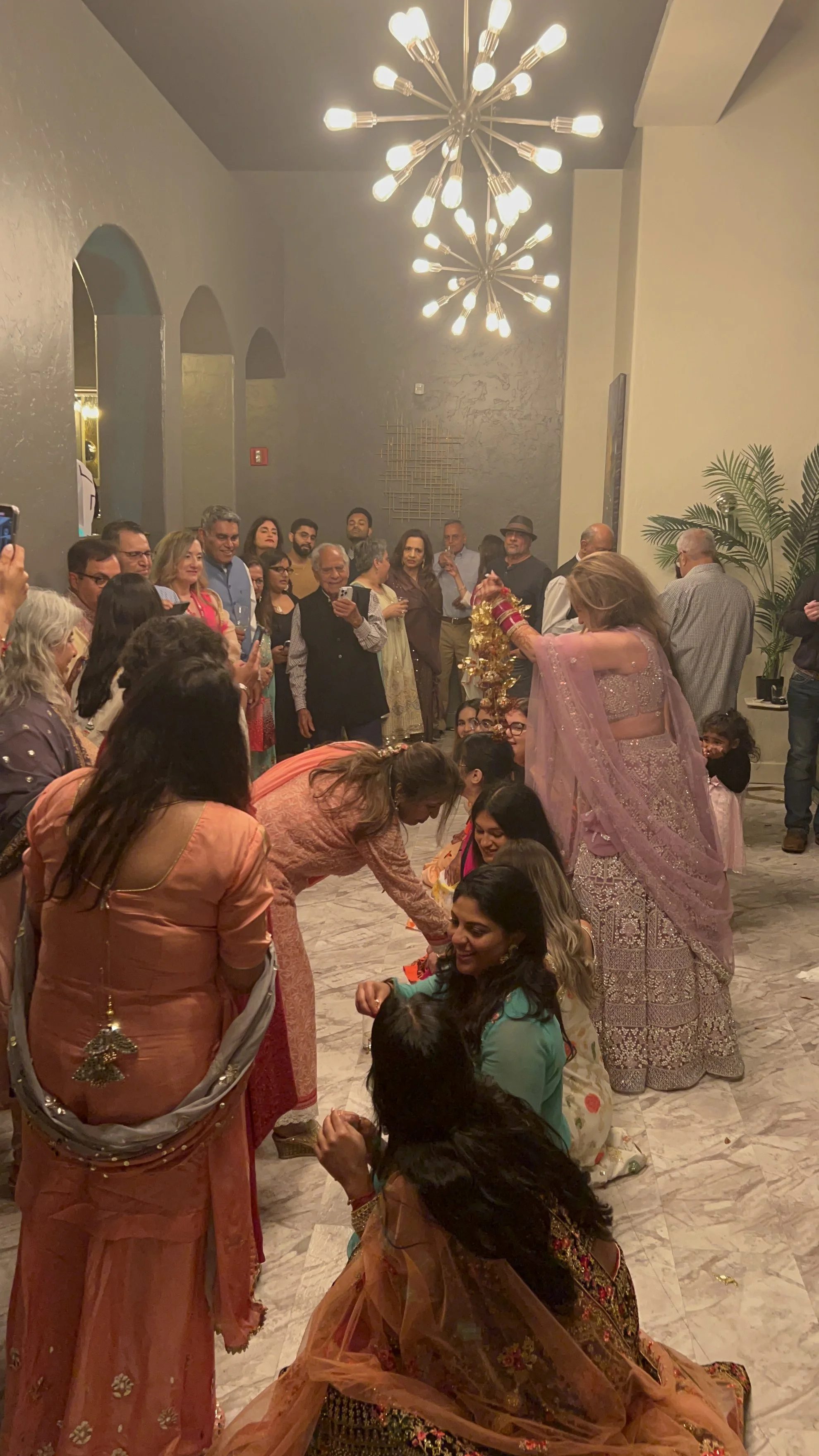 People participating in a traditional Indian ceremony, with women dressed in colorful sarees and men in formal attire, inside a decorated indoor venue with modern chandeliers and potted plants.