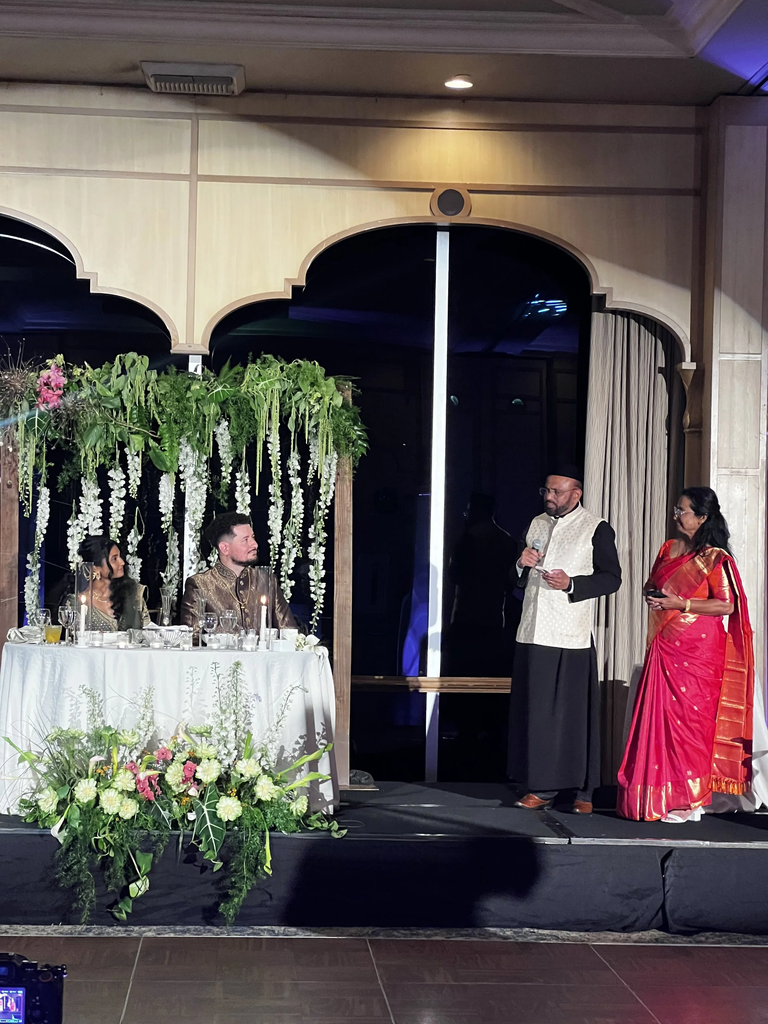Indoor event with a decorated floral backdrop, people seated at a table, and a man in traditional Indian attire speaking into a microphone while a woman in a red sari stands nearby.