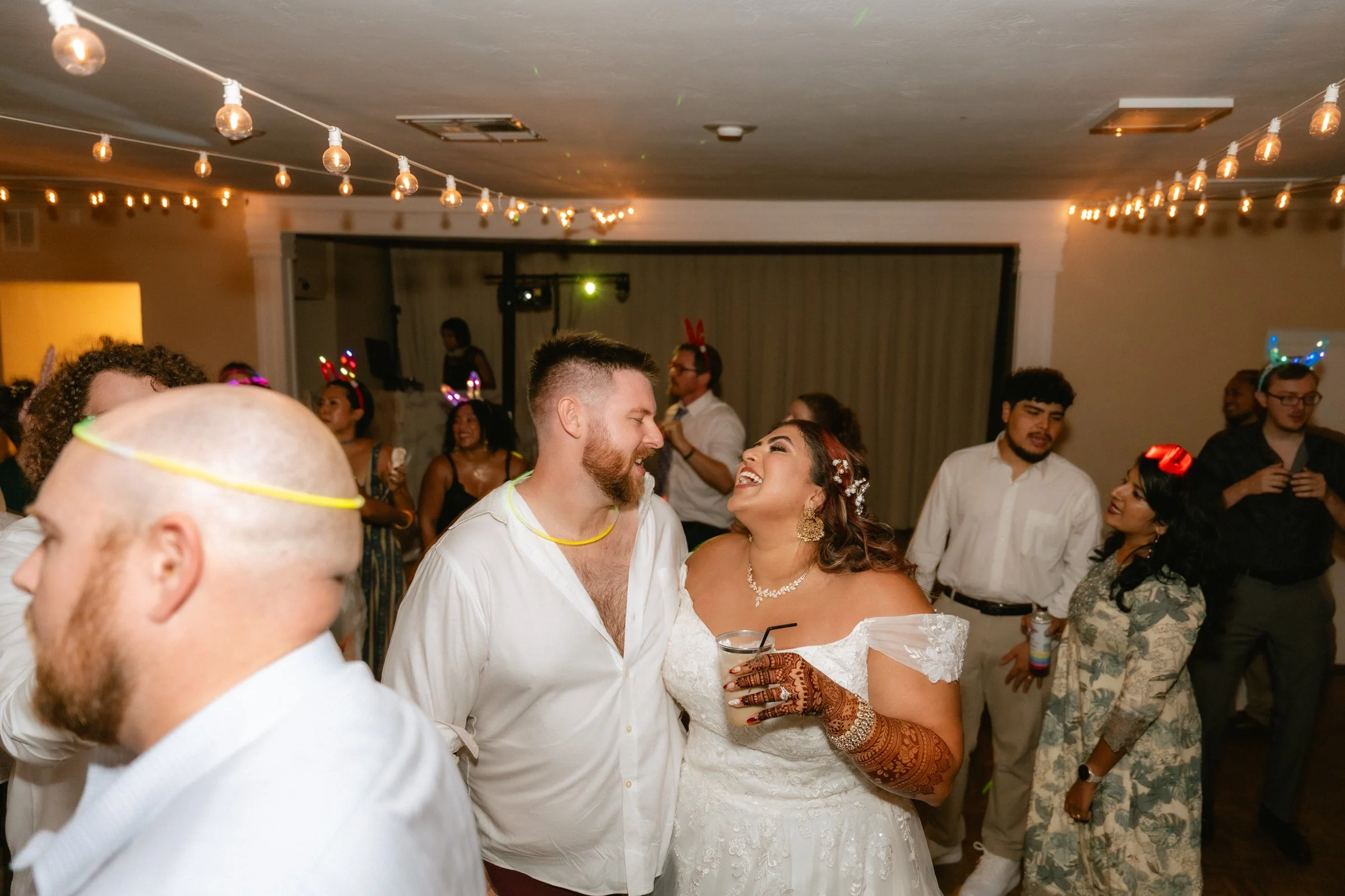 Couple dancing at a celebration or wedding reception, with friends around, festive lighting, and some guests wearing colorful headbands with lights.